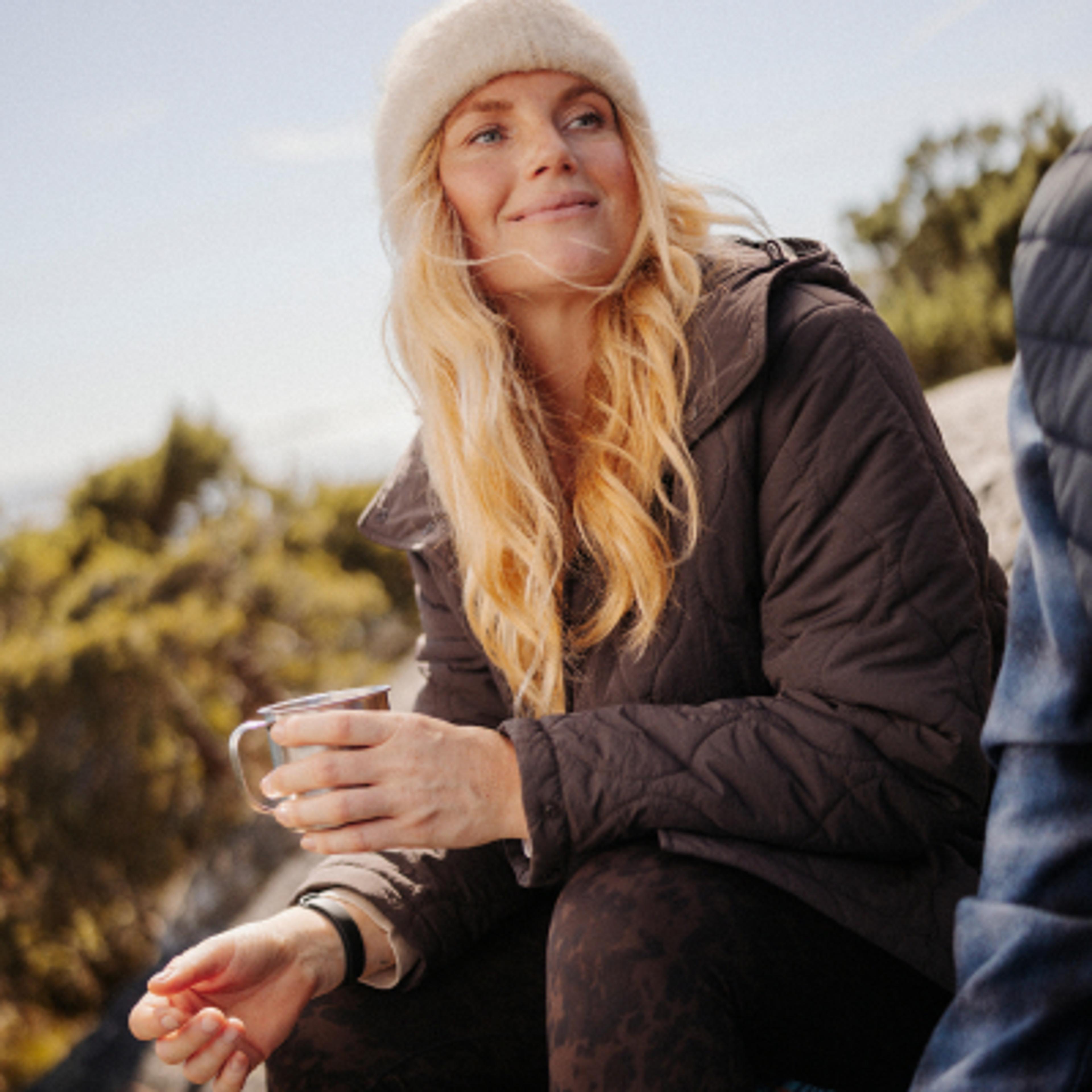 A blonde woman in a beanie and quilted jacket smiles while holding a metal mug outdoors.