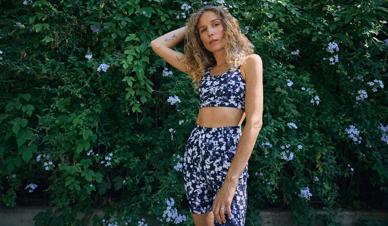A woman in a blue and white patterned athletic set stands in front of green foliage with purple flowers.