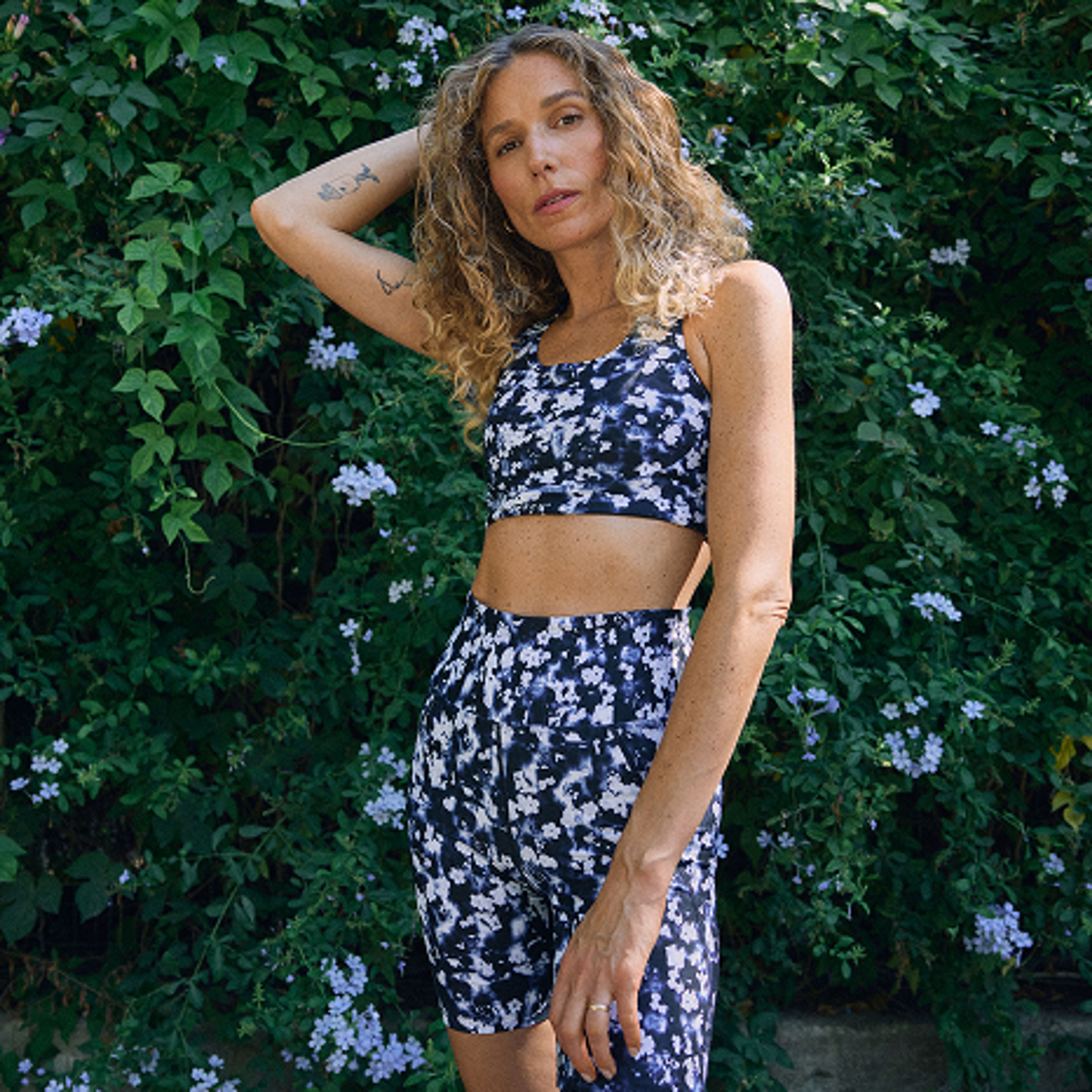 A woman in a blue and white patterned sports bra and biker shorts stands in front of a green leafy bush with small blue flowers.