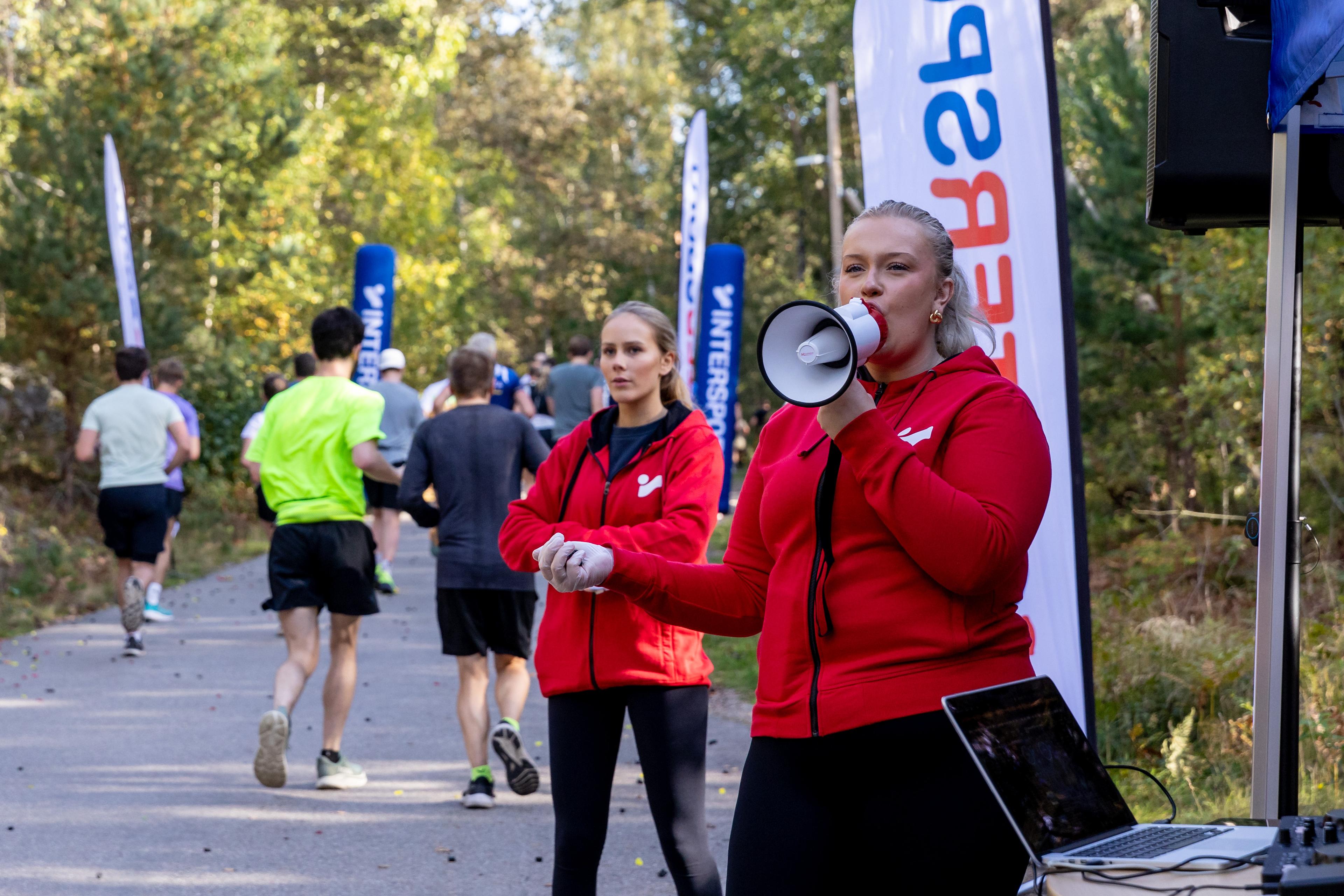 Two women in red Intersport jackets, one speaking into a megaphone, at an outdoor running event with participants in the background.