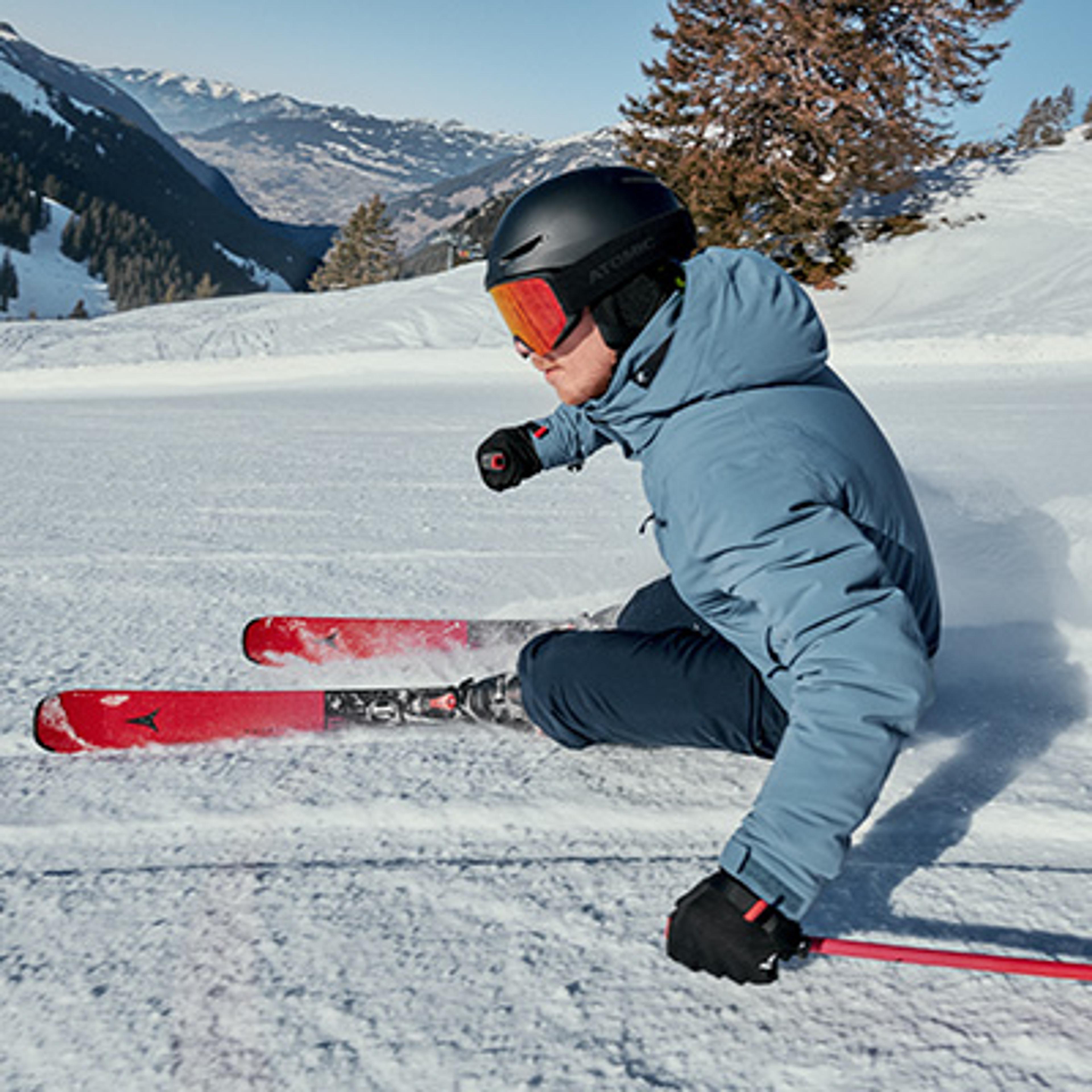A skier in a blue jacket and orange goggles carves on red skis down a snowy mountain.