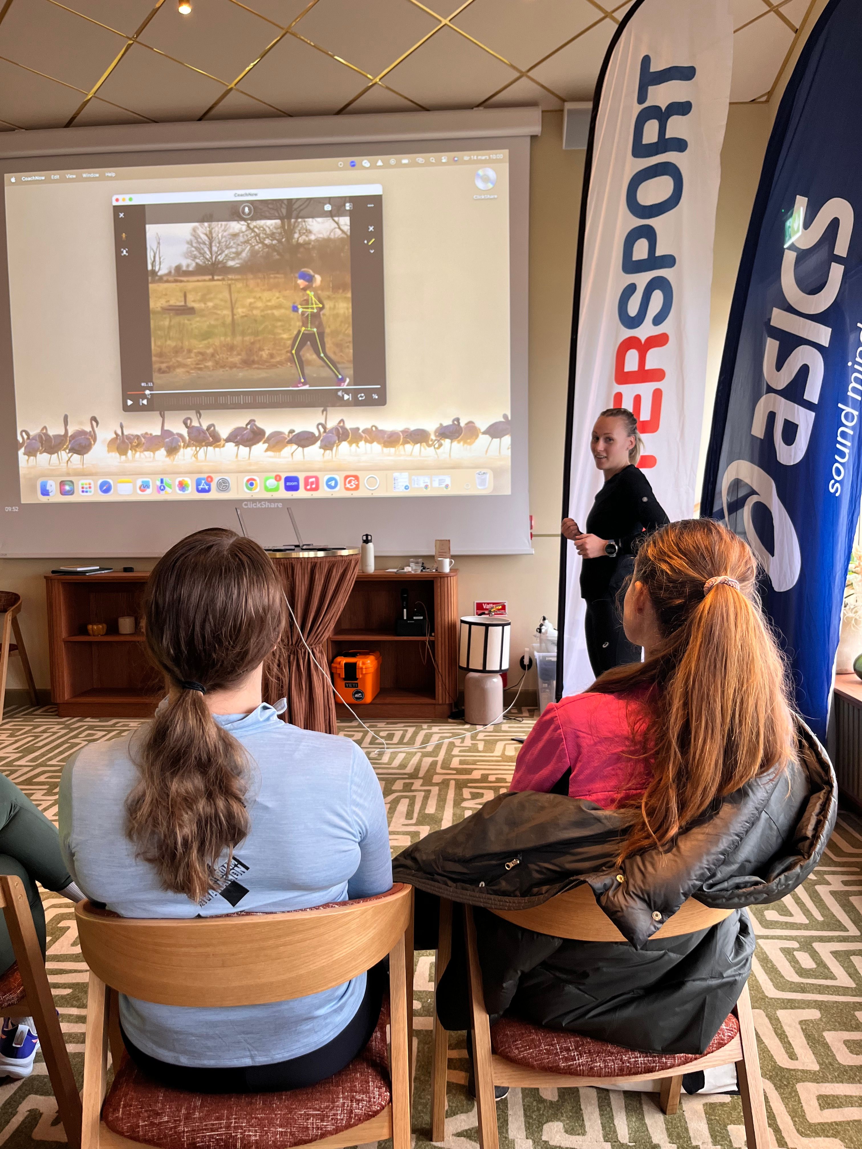 A presenter stands next to a screen showing a running video, while two attendees watch. Intersport and Asics banners are visible.