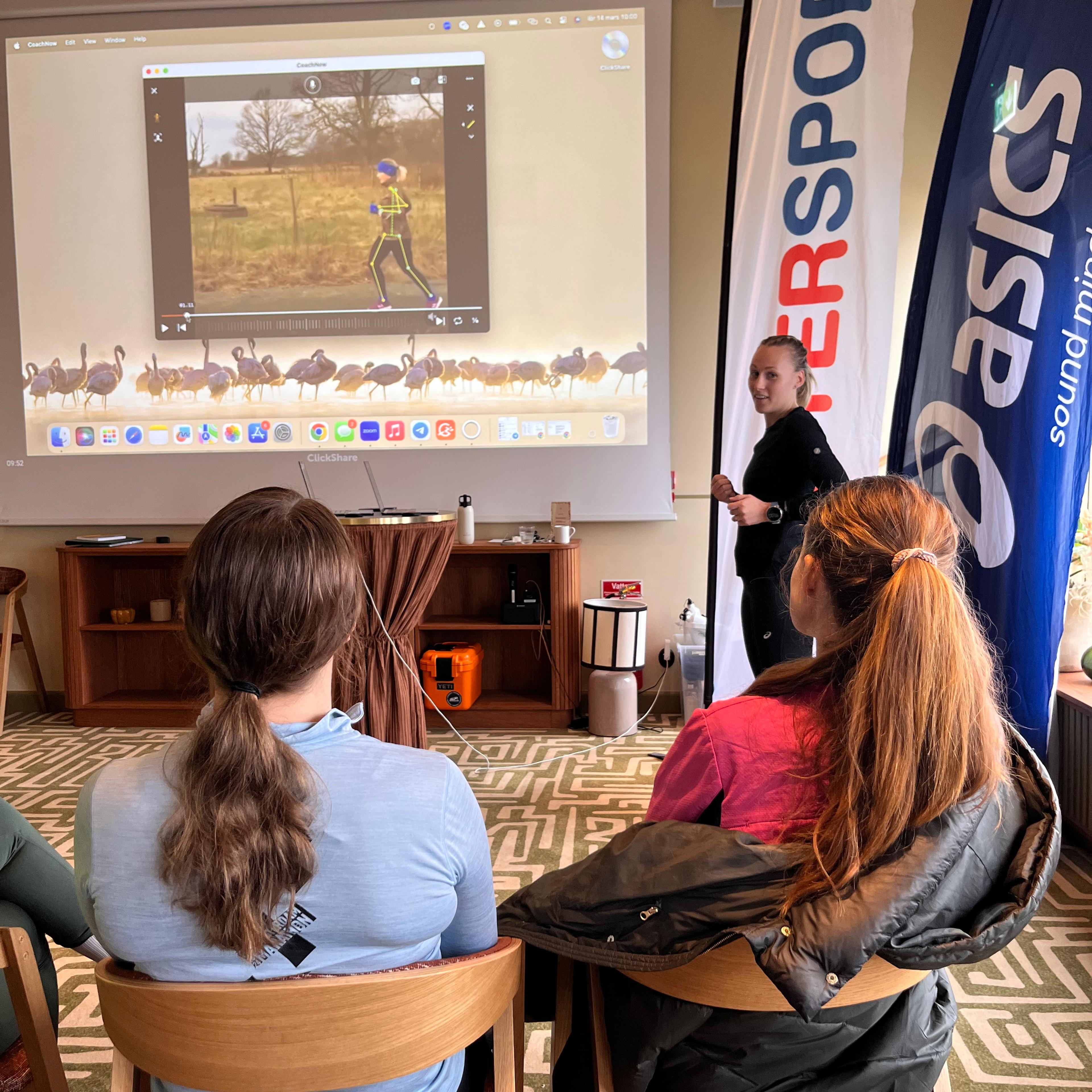 A presenter stands next to a screen showing a running video, while two attendees watch. Intersport and Asics banners are visible.