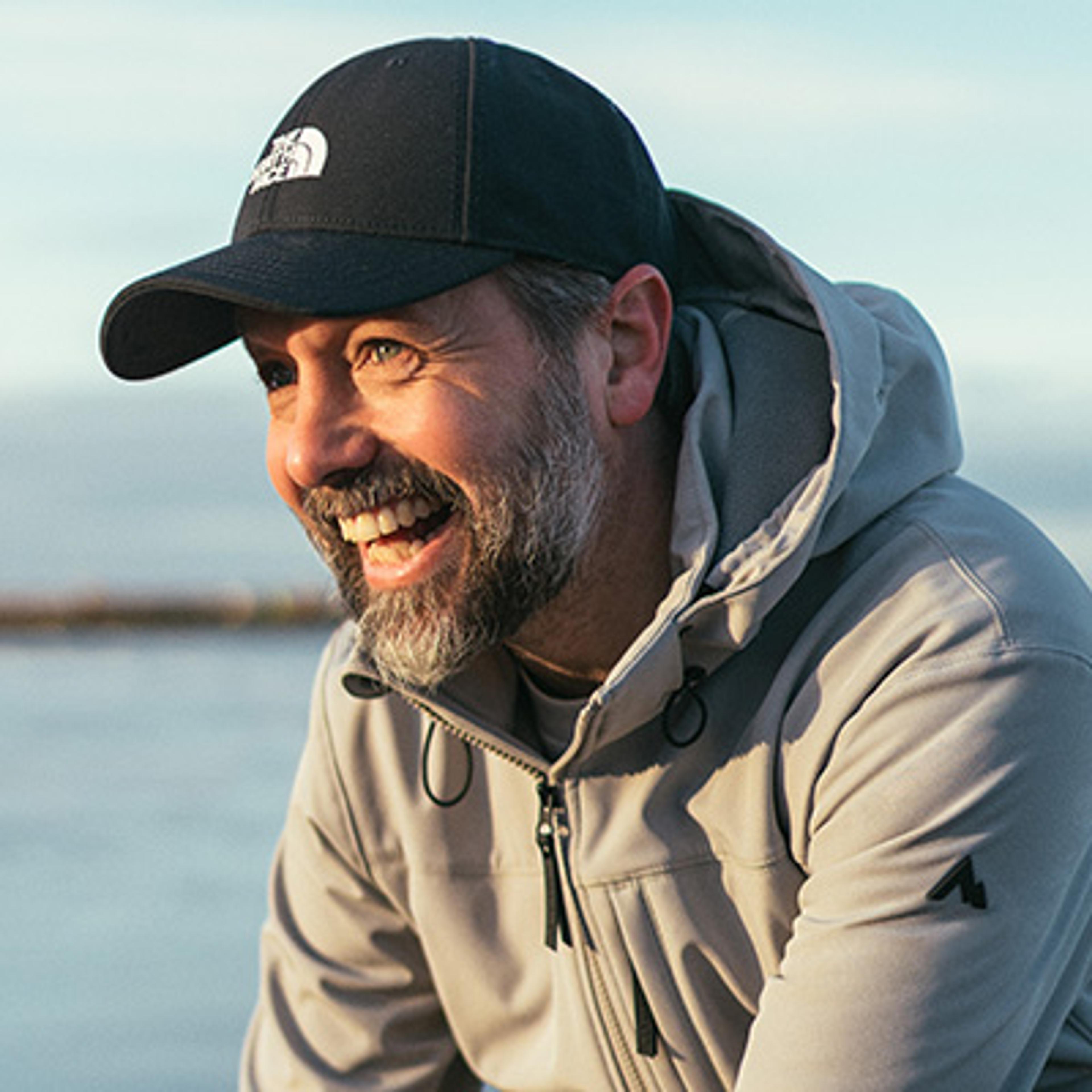 A smiling man with a beard wearing a black baseball cap and grey hooded jacket.