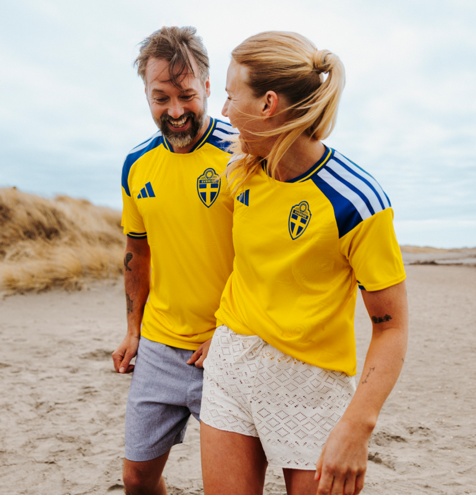 A smiling man and woman wearing yellow Swedish football jerseys on a sandy beach.