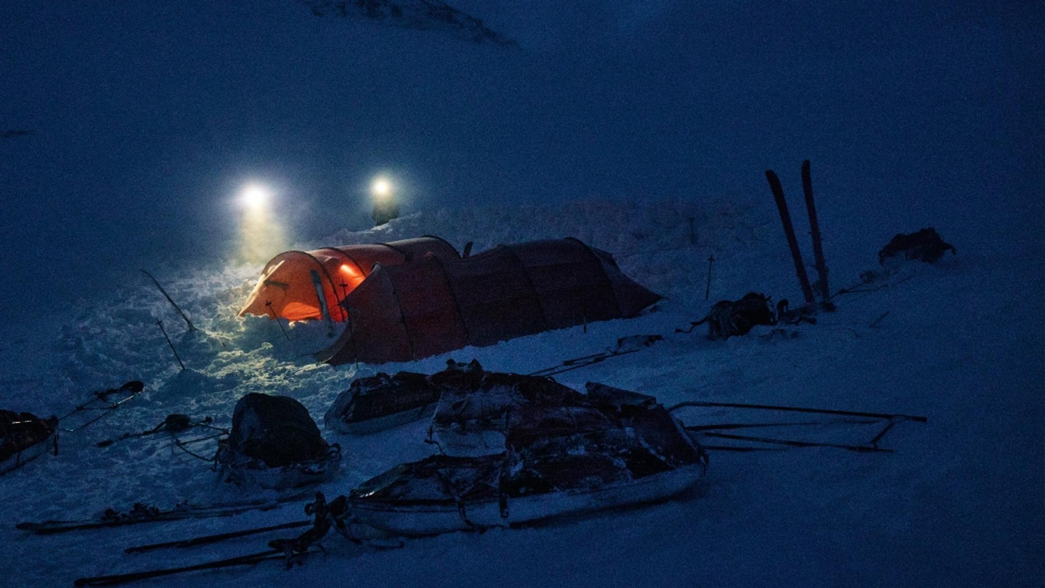 A winter expedition camp at night with an illuminated orange tent, skis, and equipment on snow.