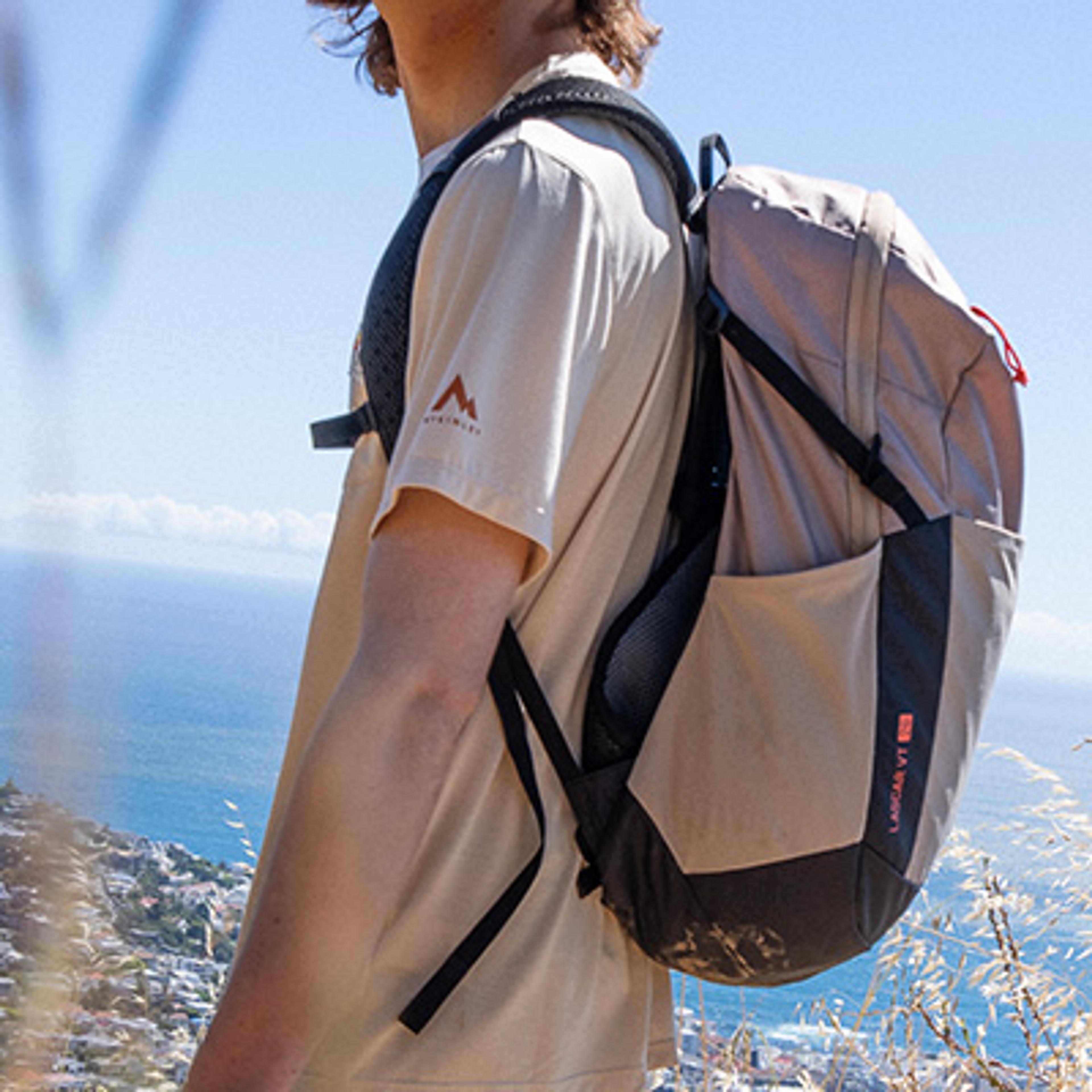 Hiker wearing a beige t-shirt and a two-tone beige and black "LANCAR VT 10" backpack, overlooking a blue ocean and a coastal town.