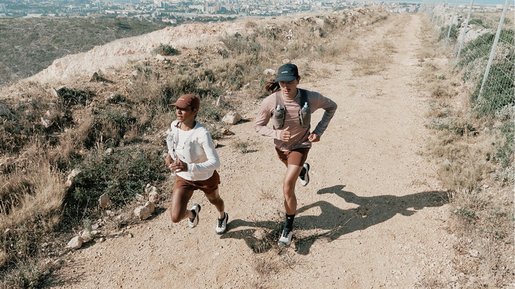 Two trail runners on a dusty path with dry hills and a distant city.
