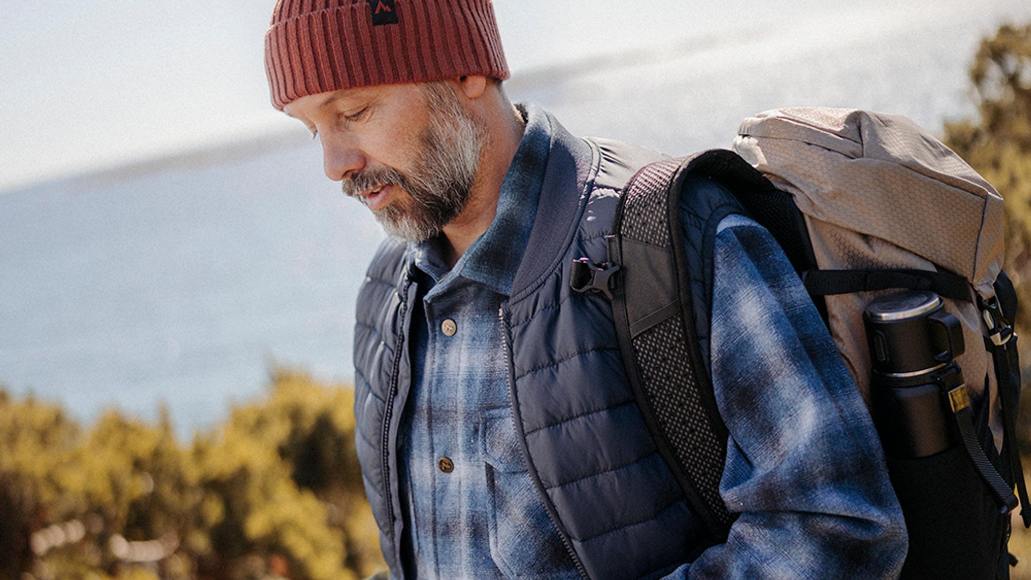 Hiker in a beanie and vest carrying a backpack with a thermos, by water.