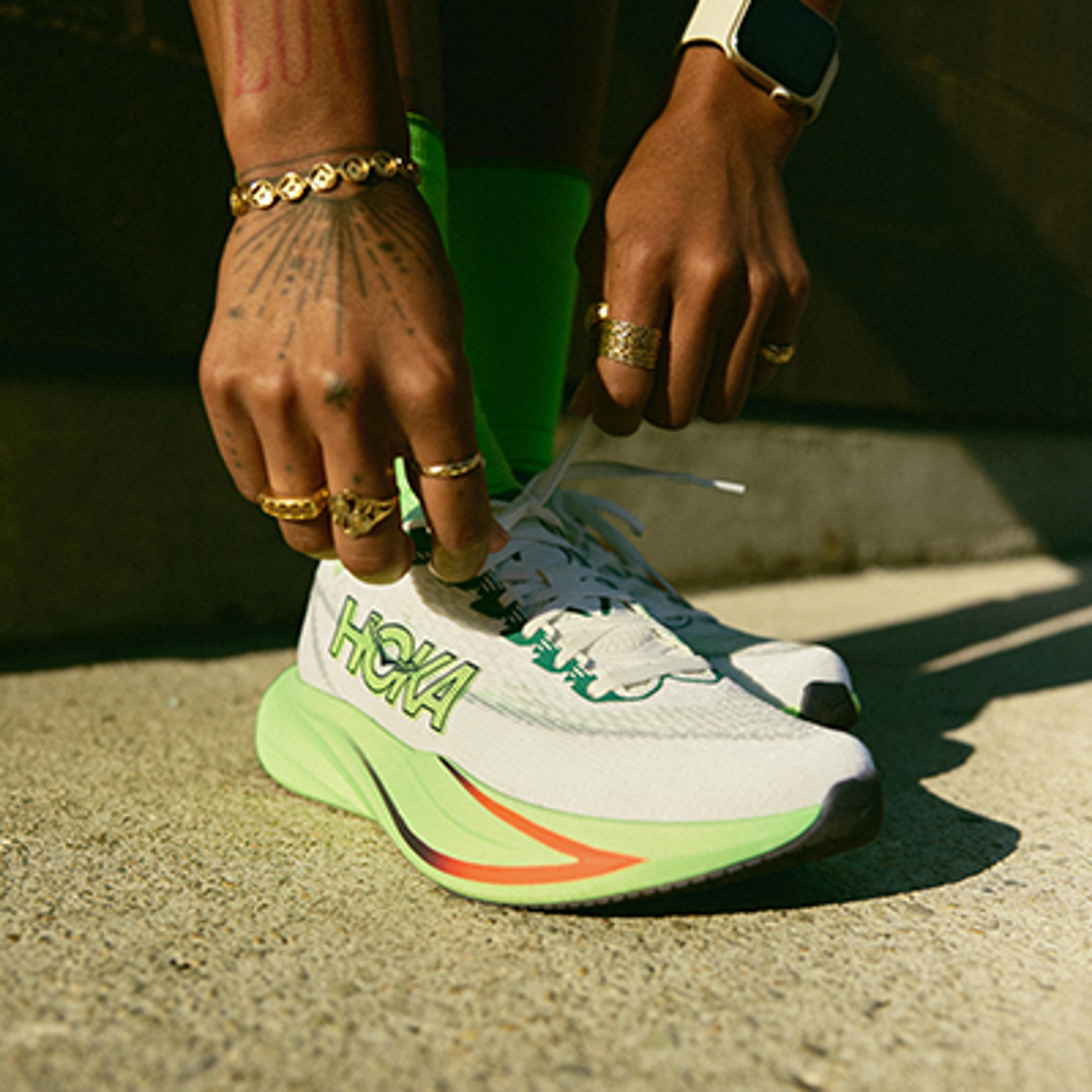 Hands adorned with gold rings tie the laces of a white Hoka running shoe with neon green and orange accents.