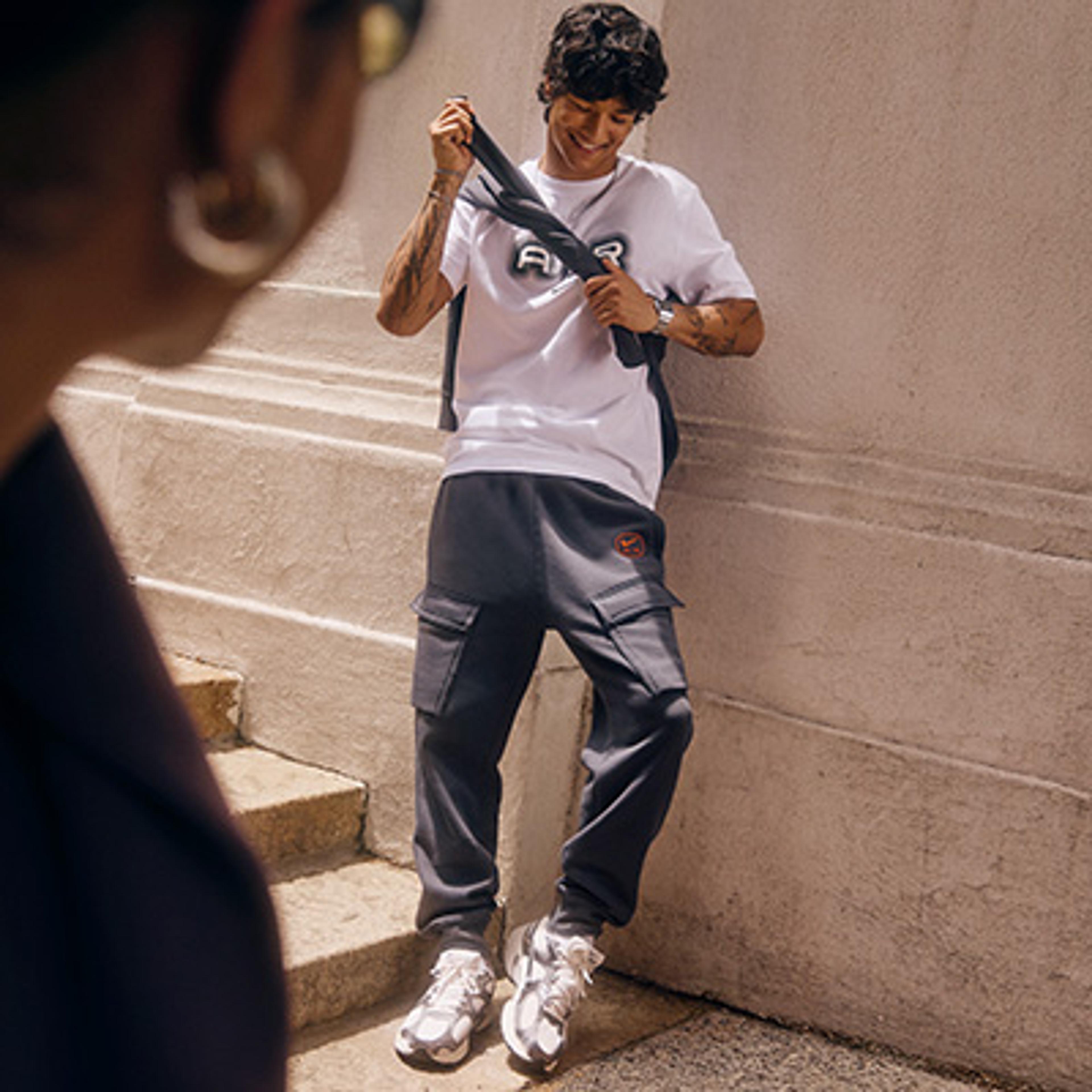 A smiling man in a white "AIR" t-shirt, dark cargo joggers, and white sneakers leans against a wall.