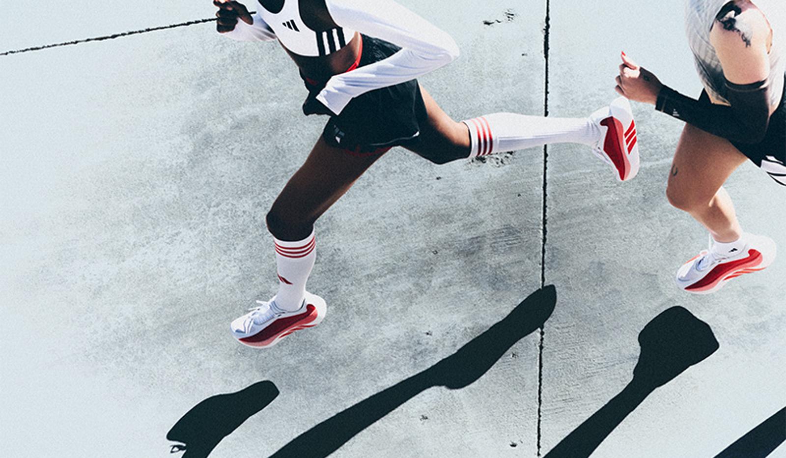 Overhead view of two runners wearing white and red athletic shoes and socks, casting long shadows on a concrete surface.