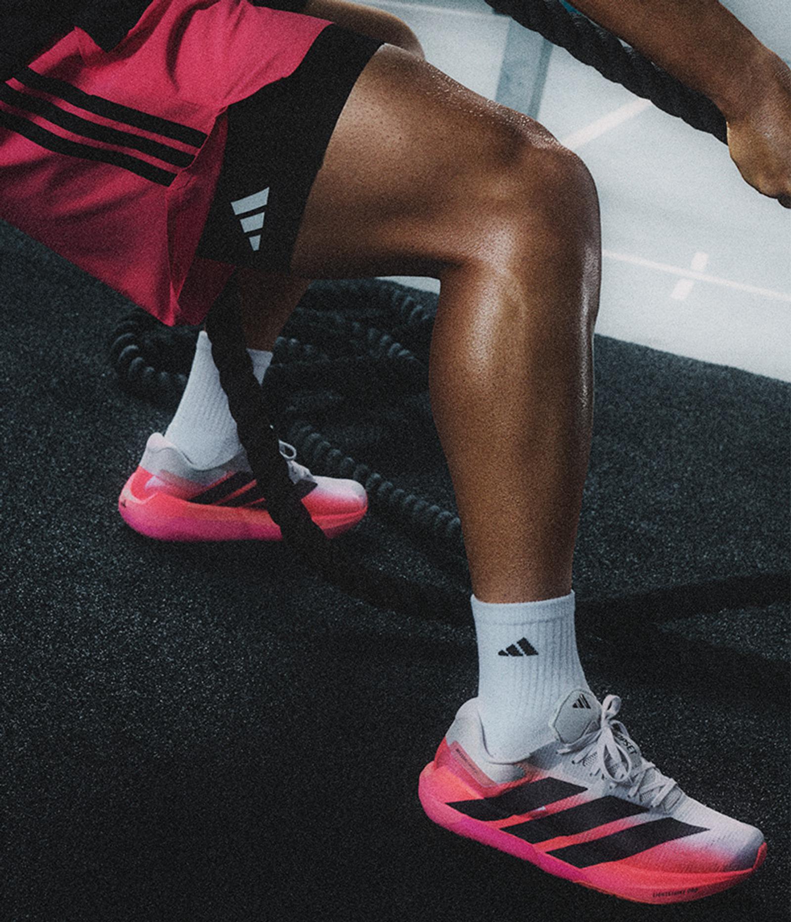 A person's sweaty legs in pink shorts and white-pink Adidas running shoes, gripping a battle rope.