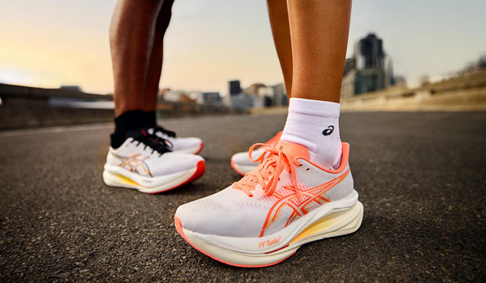 Close-up of two pairs of white and orange ASICS running shoes on asphalt, with a blurred cityscape behind them.