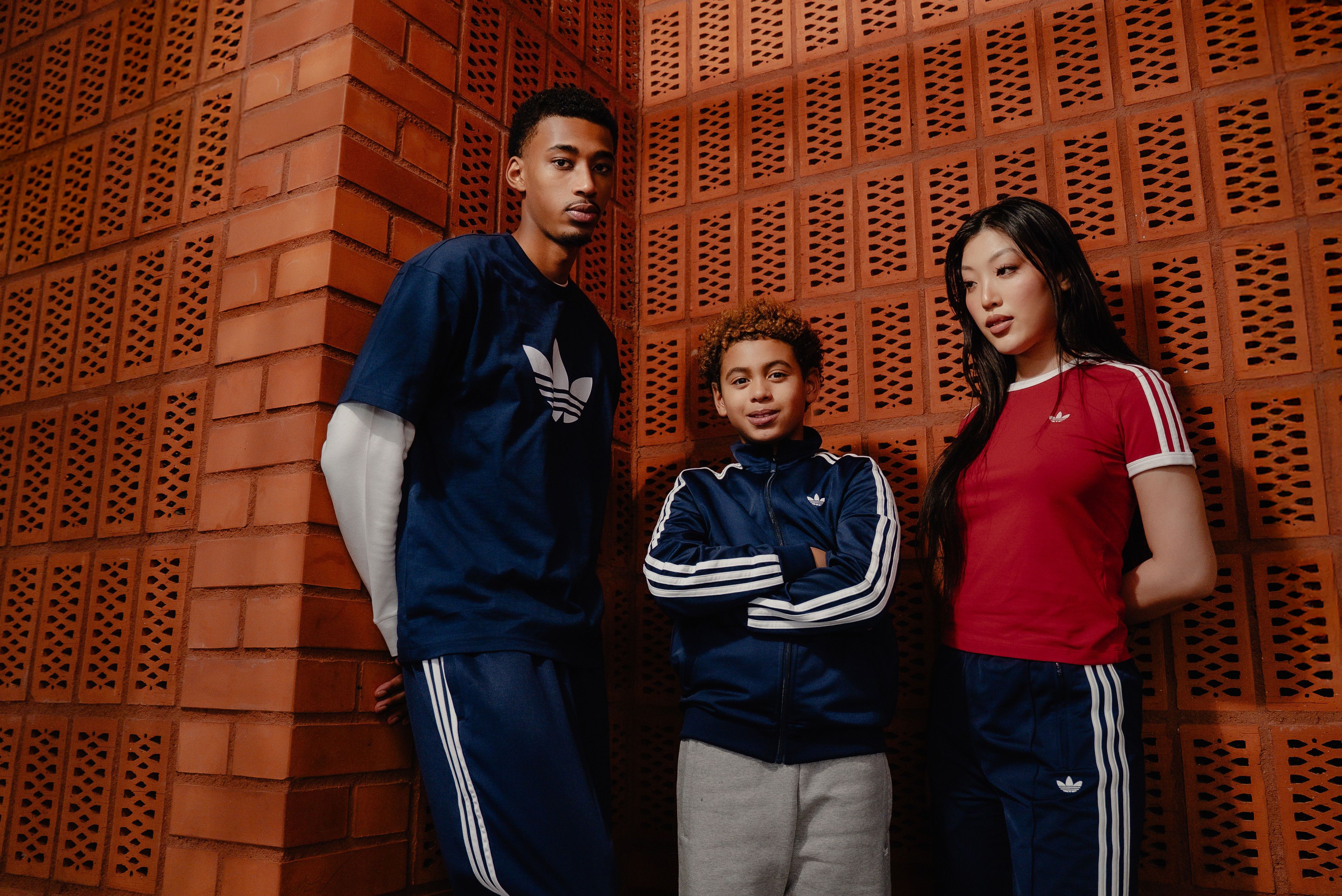 A man, a child, and a woman wearing Adidas sportswear stand against a brick wall.
