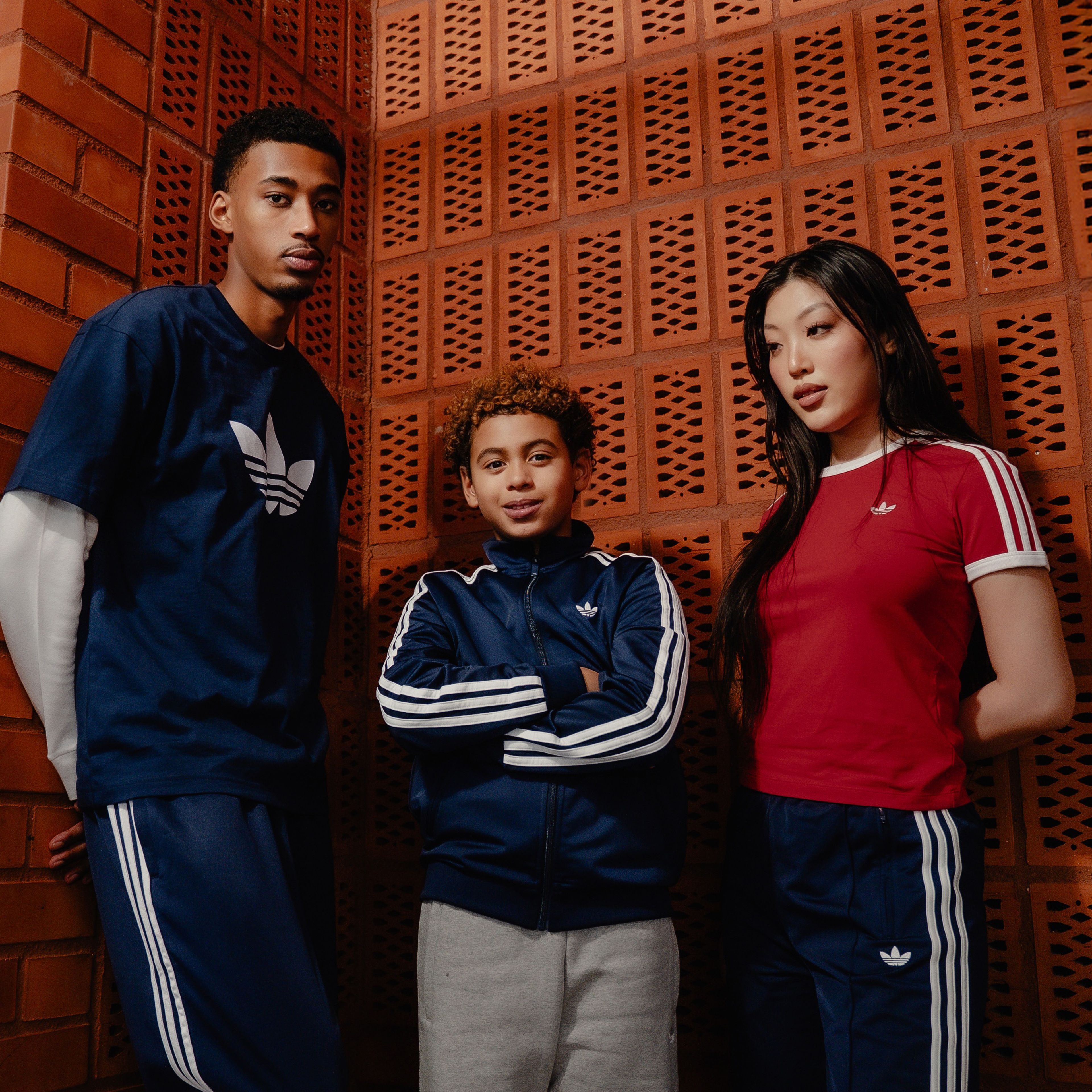 A man, a child, and a woman wearing Adidas sportswear stand against a brick wall.