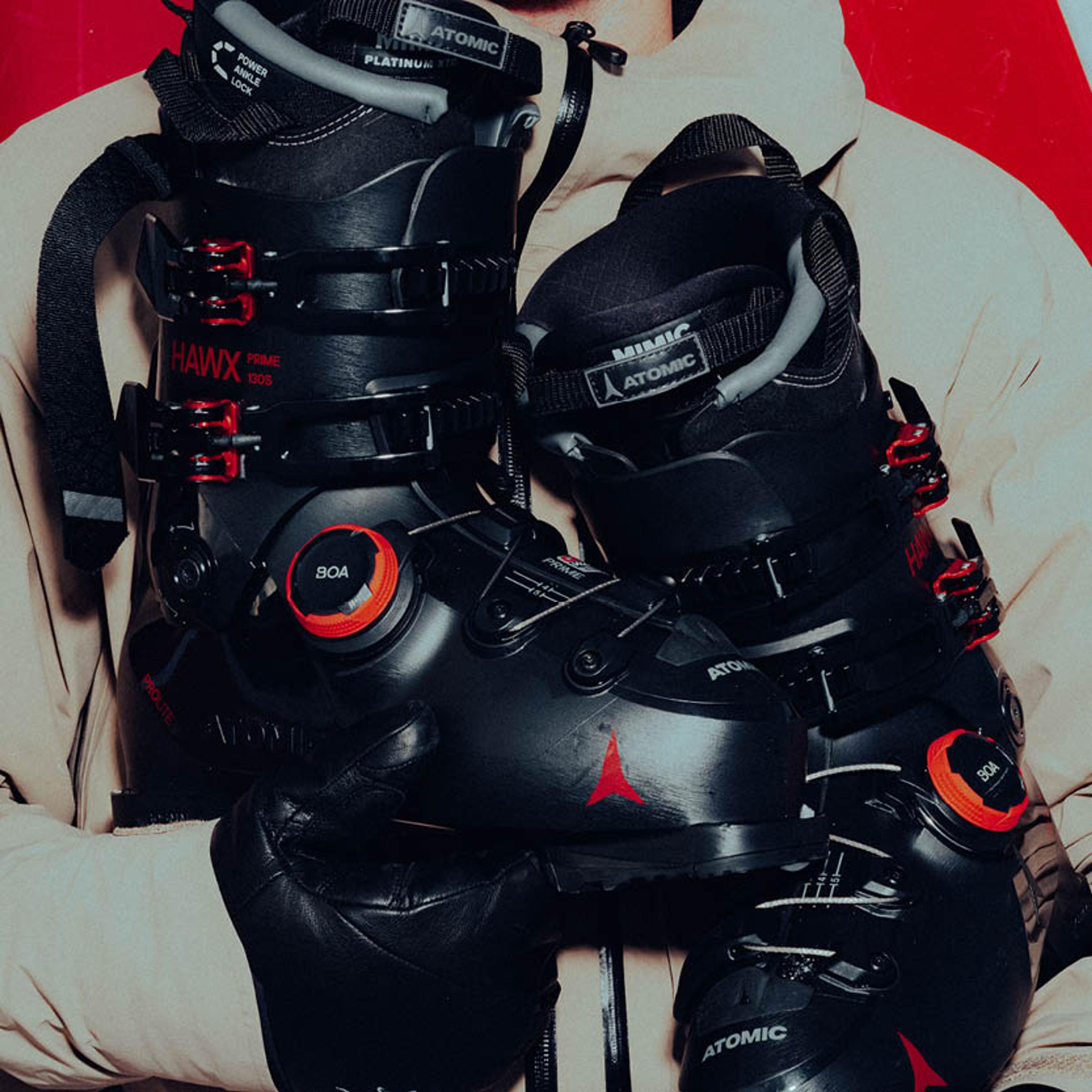 Person in a tan jacket and black gloves holds two black Atomic ski boots with red accents.