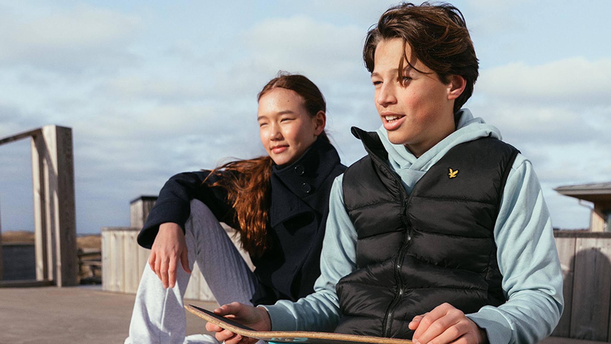 A boy holding a skateboard and a girl sitting next to him outdoors on a sunny day.