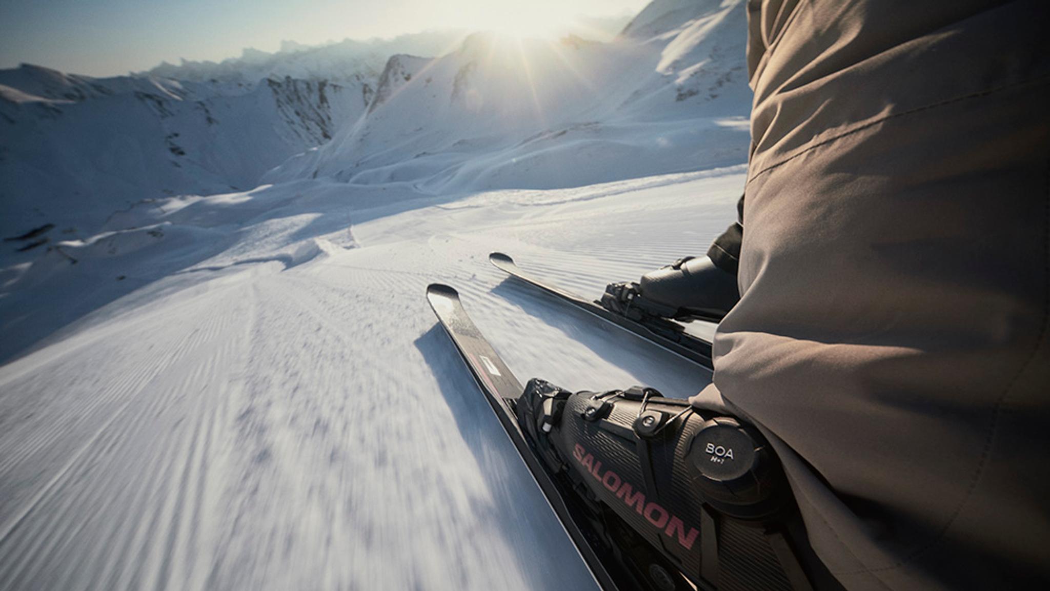 A skier's first-person view looking down a bright, snowy mountain slope.