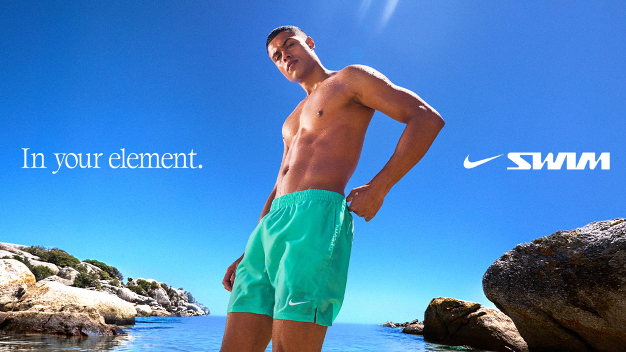 A man in green Nike swim shorts on a sunny beach with text "In your element." and "Nike SWIM".