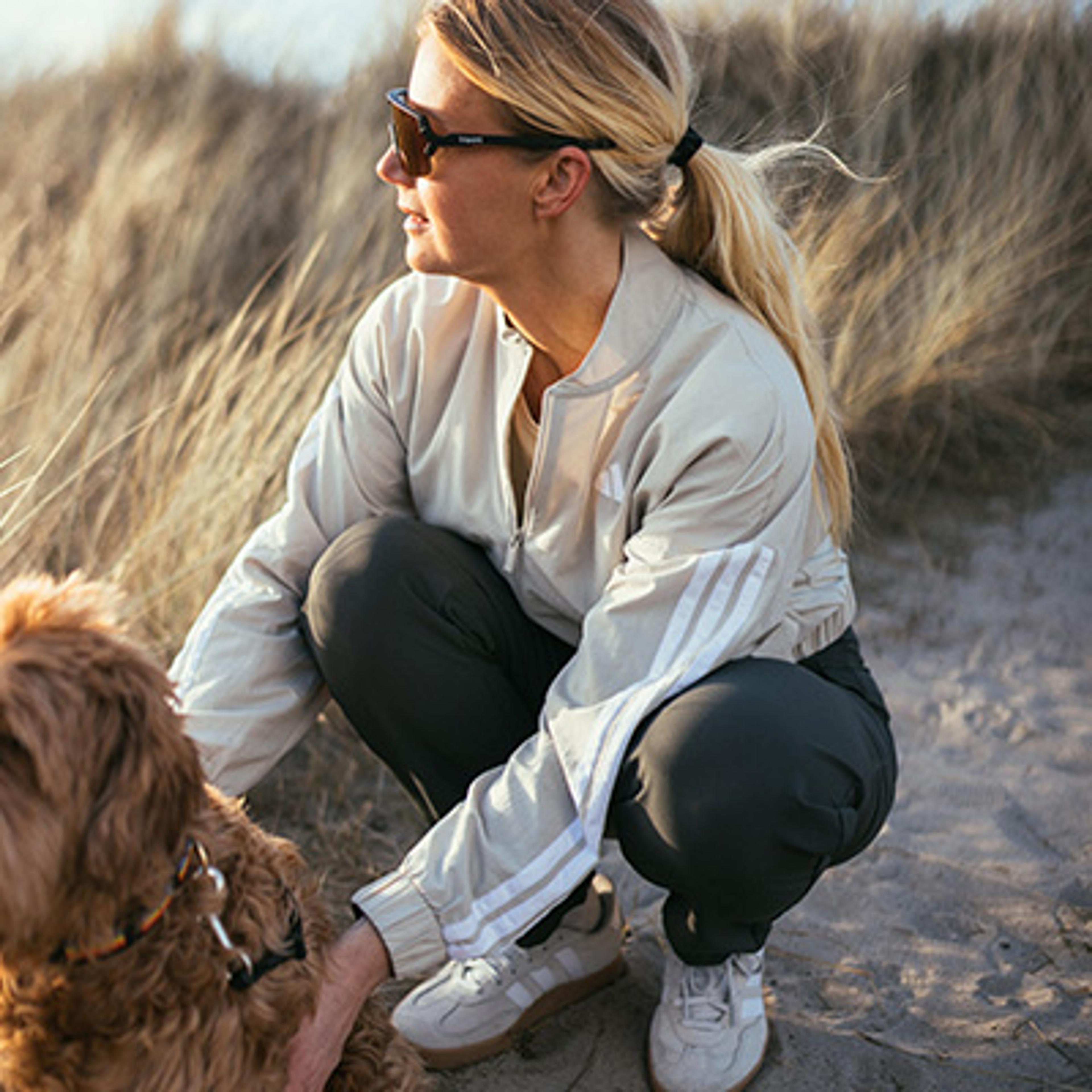 A woman in sunglasses crouches to pet a brown dog on a sandy patch with dry grass.