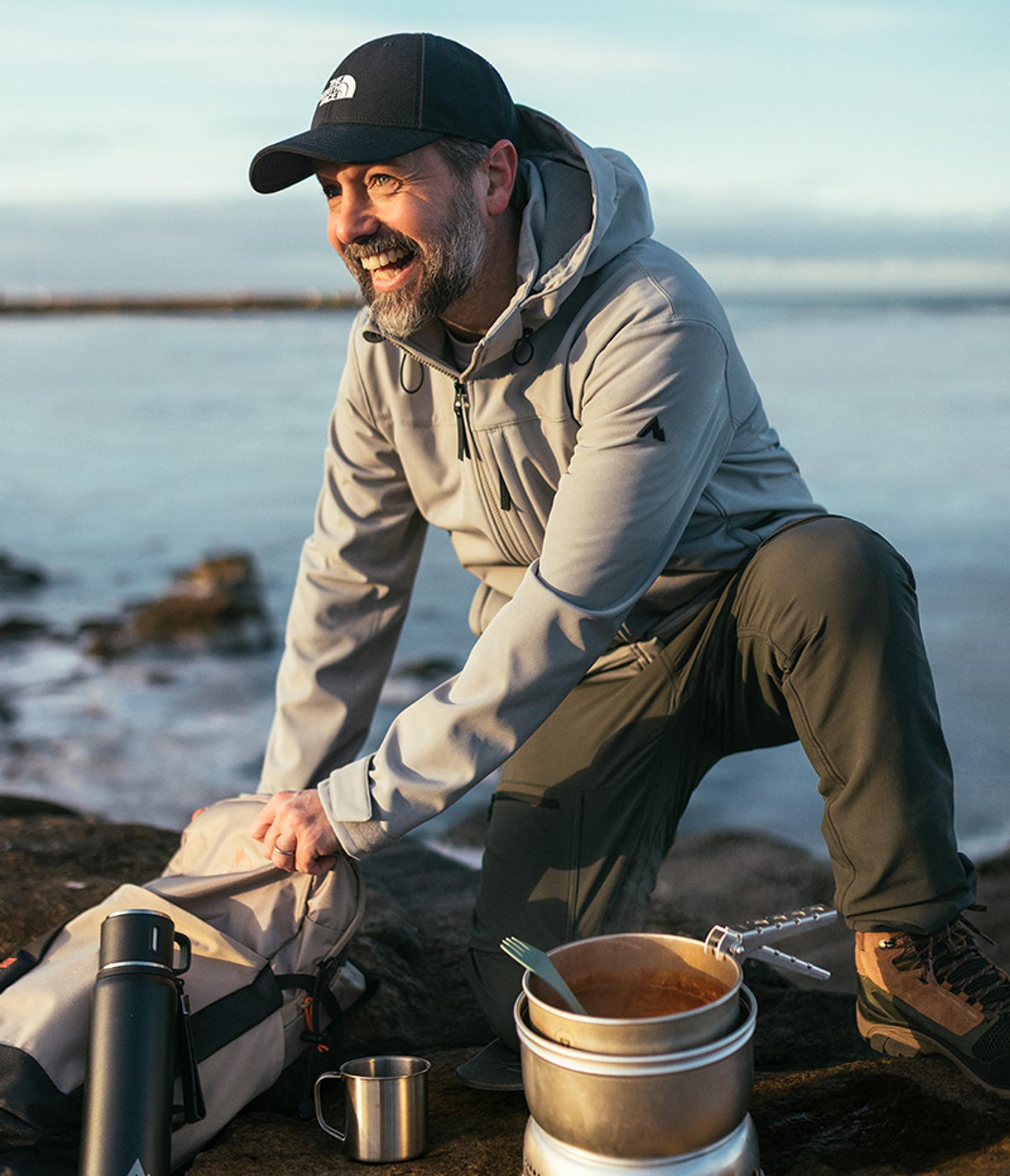 A smiling bearded man in outdoor clothing kneels by the water, opening his backpack beside a camping stove with food.