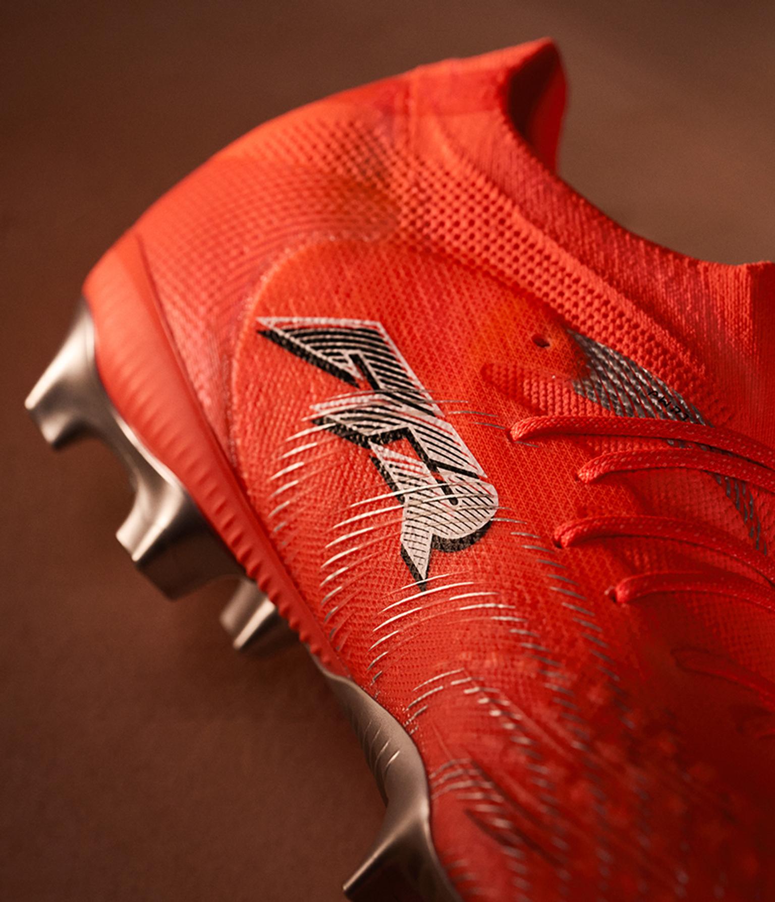 Close-up of a bright orange football boot with a stylized silver and black R logo and metallic studs.