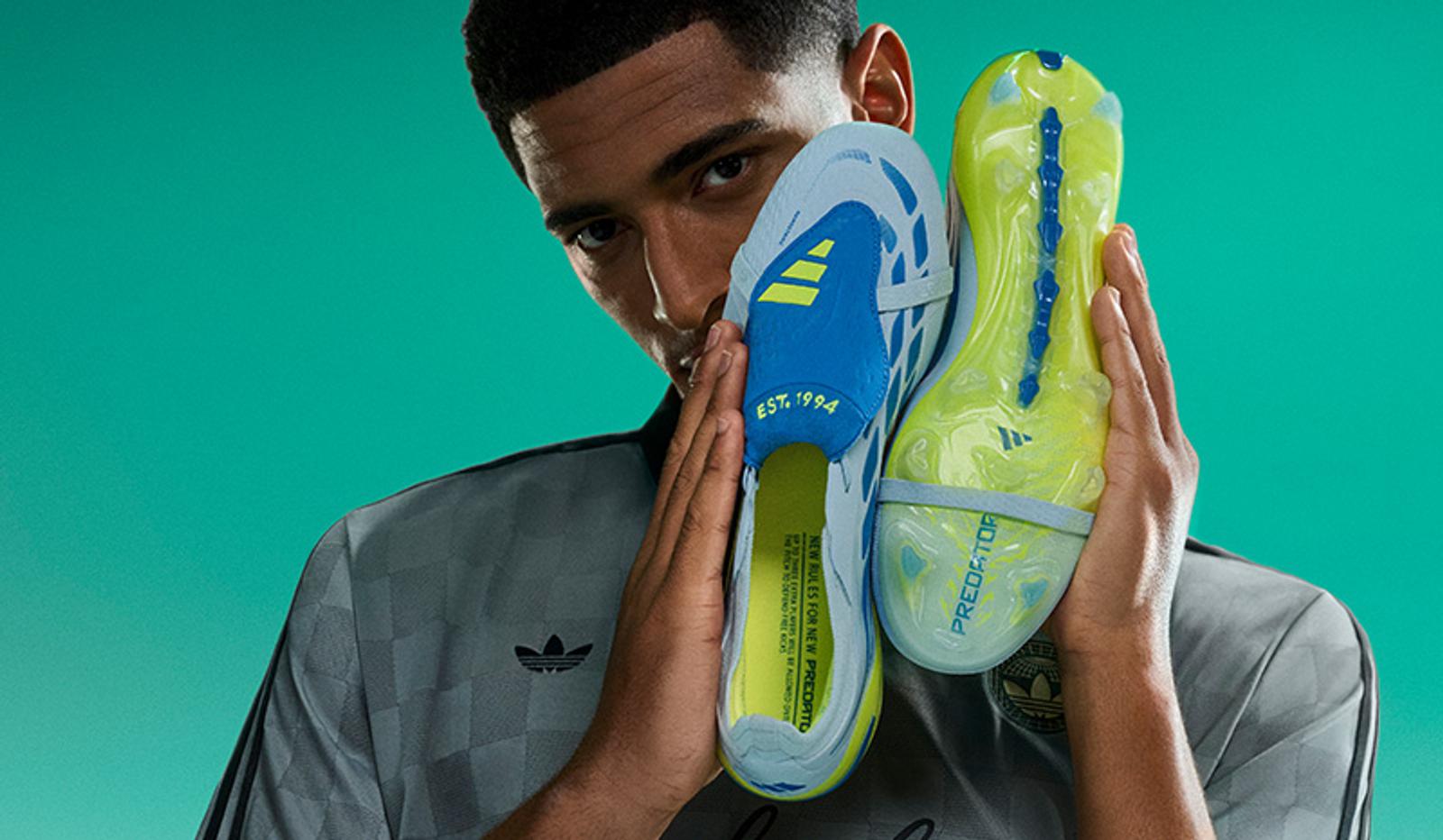 A man in an Adidas shirt holds up two light blue and neon yellow Adidas Predator soccer cleats against a teal background.