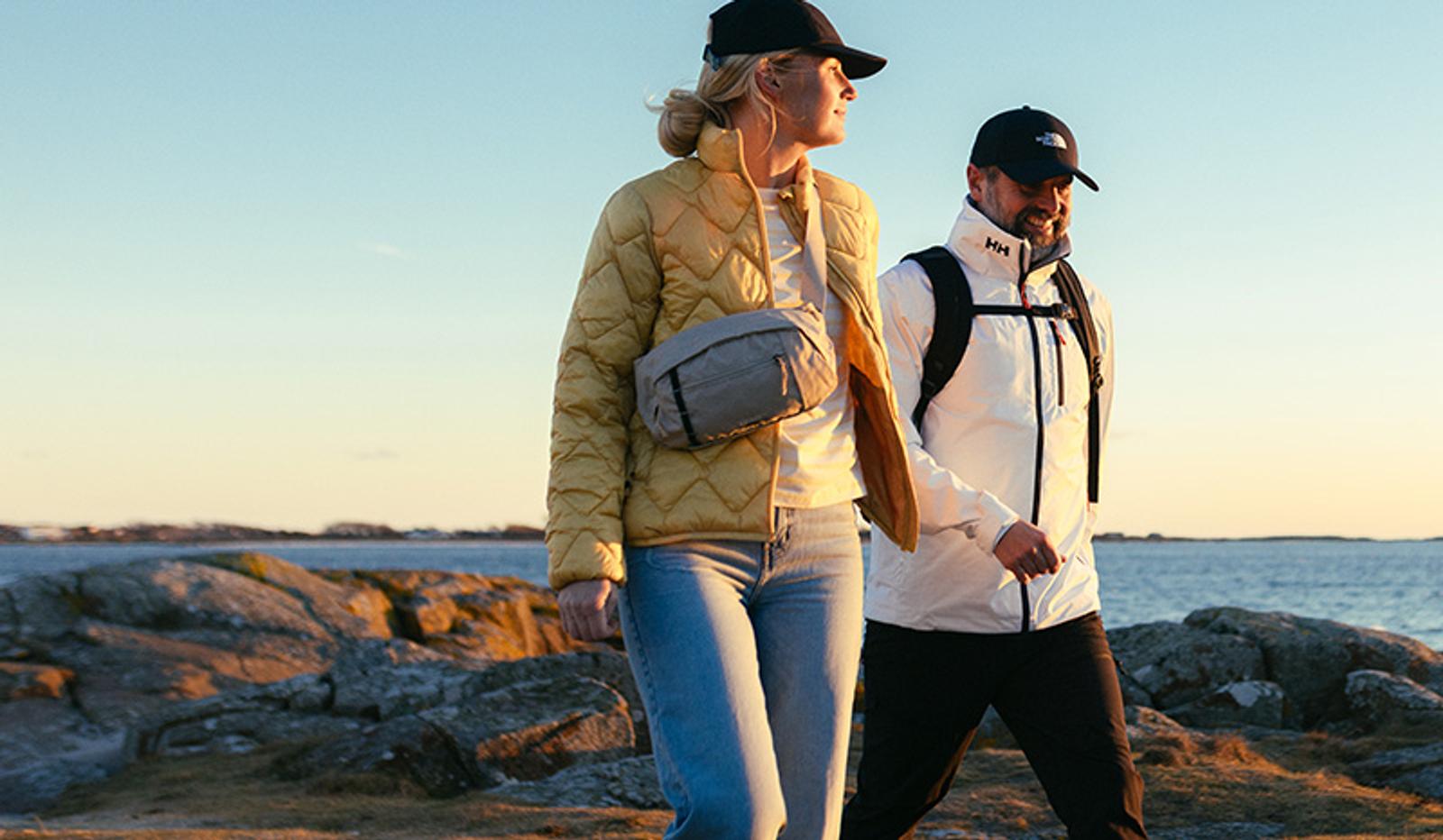 A woman in a yellow quilted jacket and a man in a white Helly Hansen jacket walk along a rocky coastline at sunset.