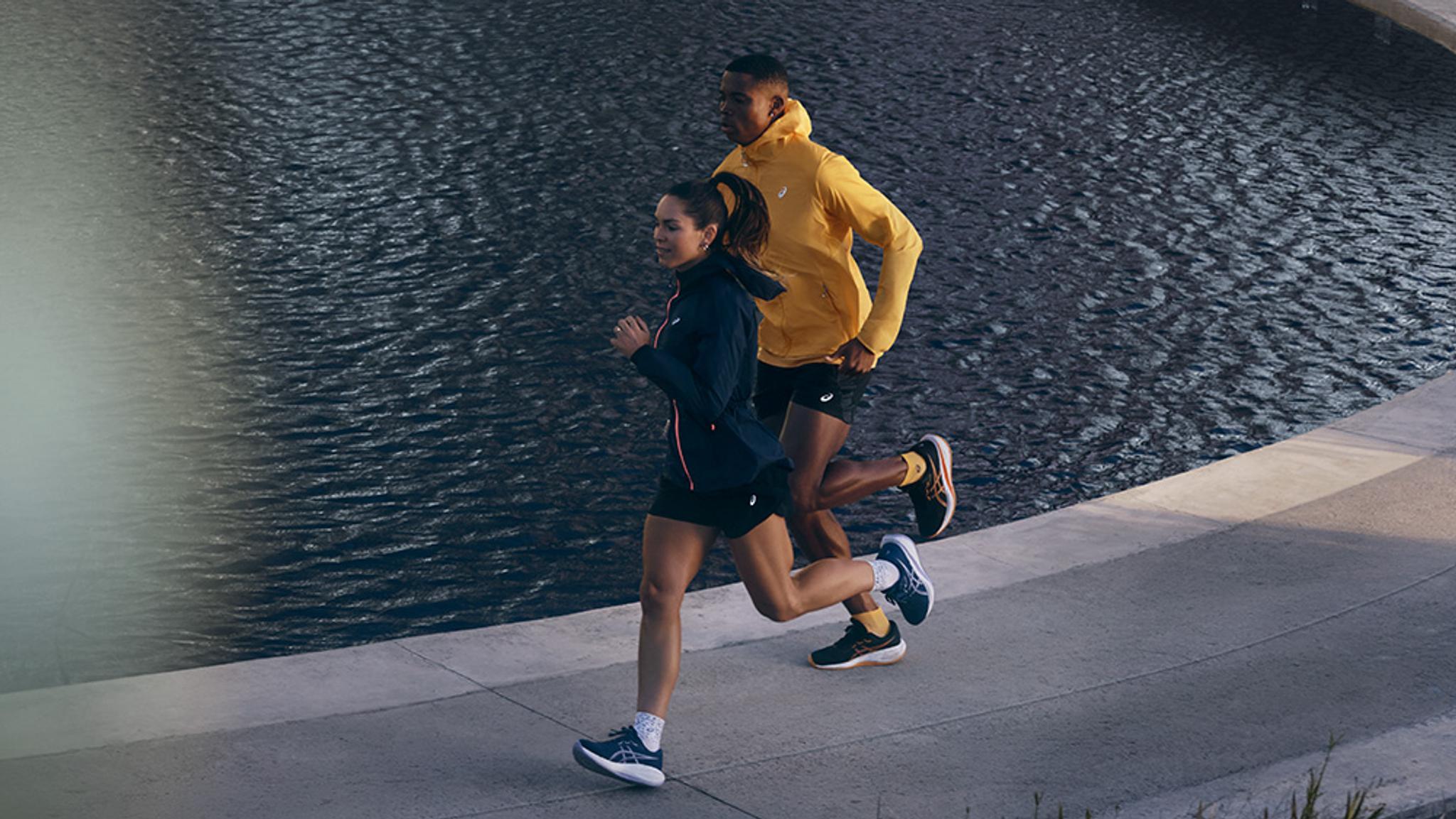 A woman in a dark jacket and a man in a yellow jacket running along a path next to water.