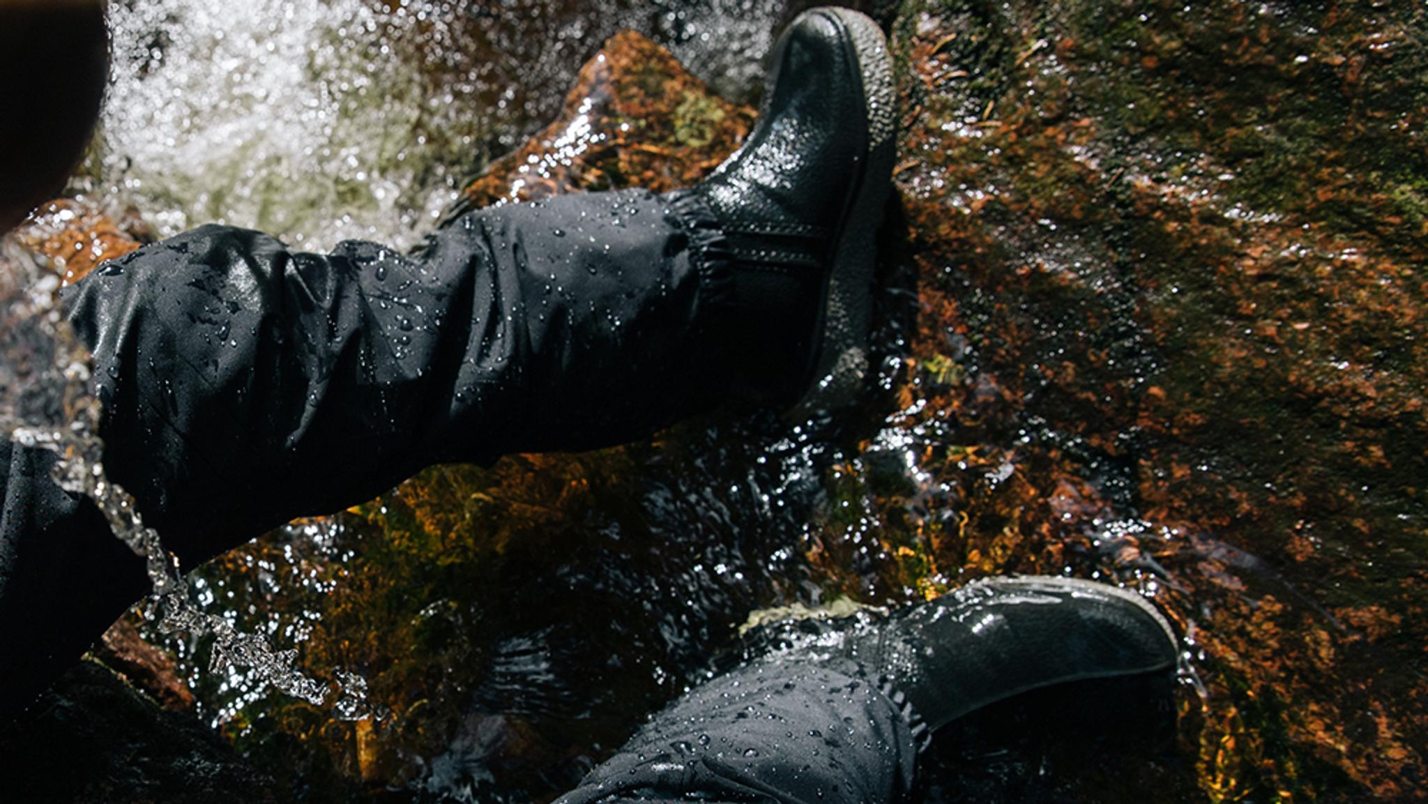 a kid wearing black boots is standing on a rock in the water .