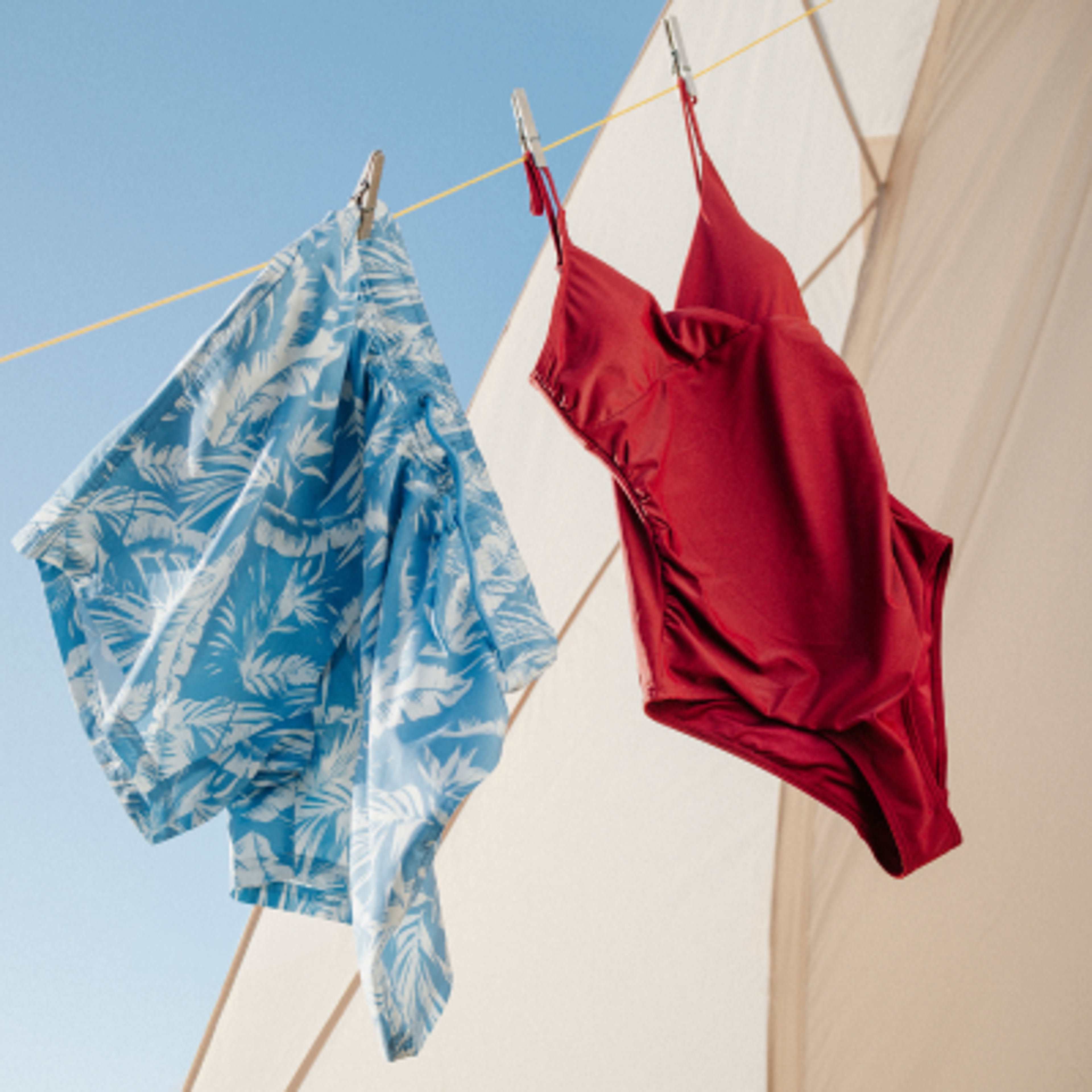 A blue tropical-print swim trunk and a red one-piece swimsuit hang on a clothesline against a blue sky and a white sail.