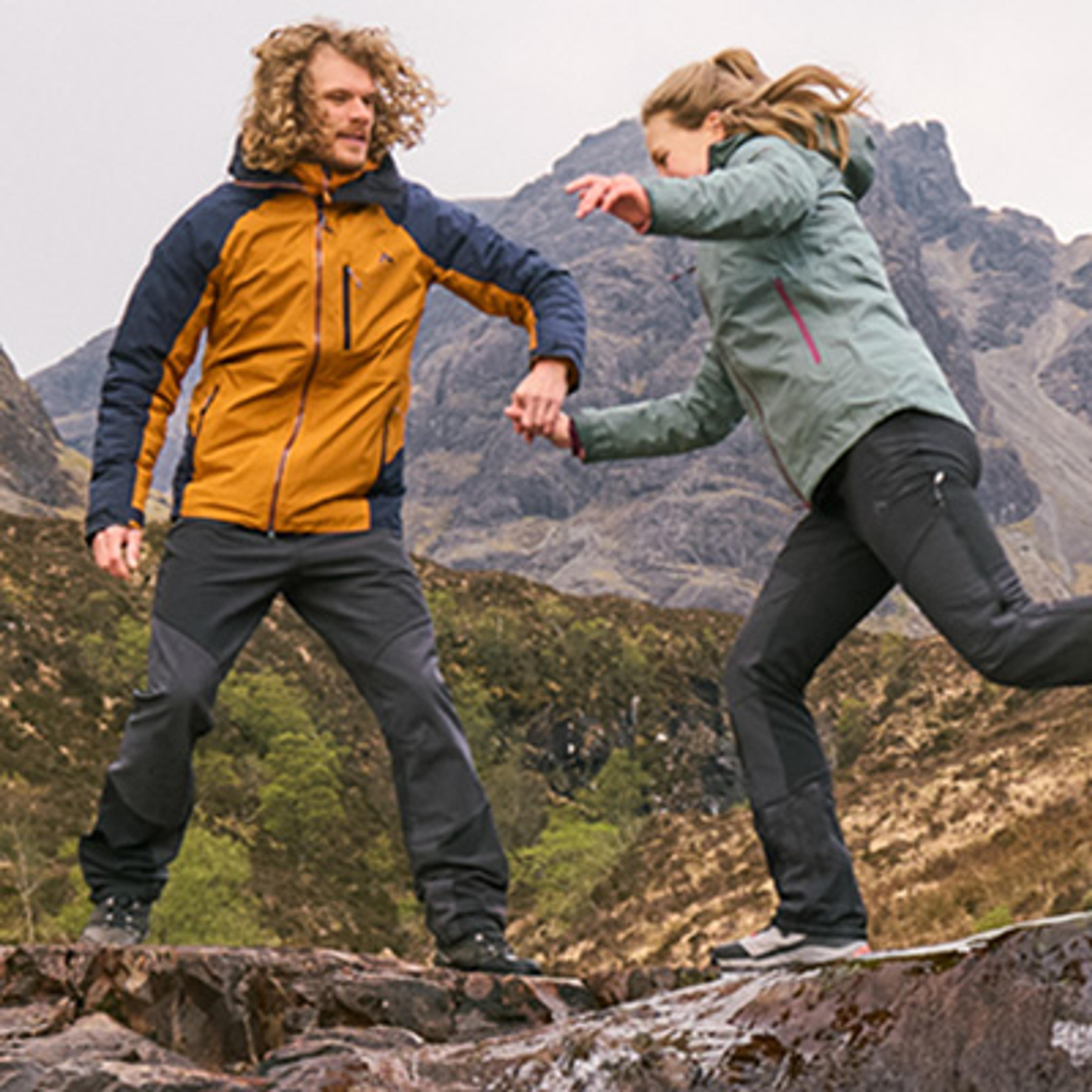 Two hikers in outdoor gear hold hands while leaping across rocks in a mountainous landscape.