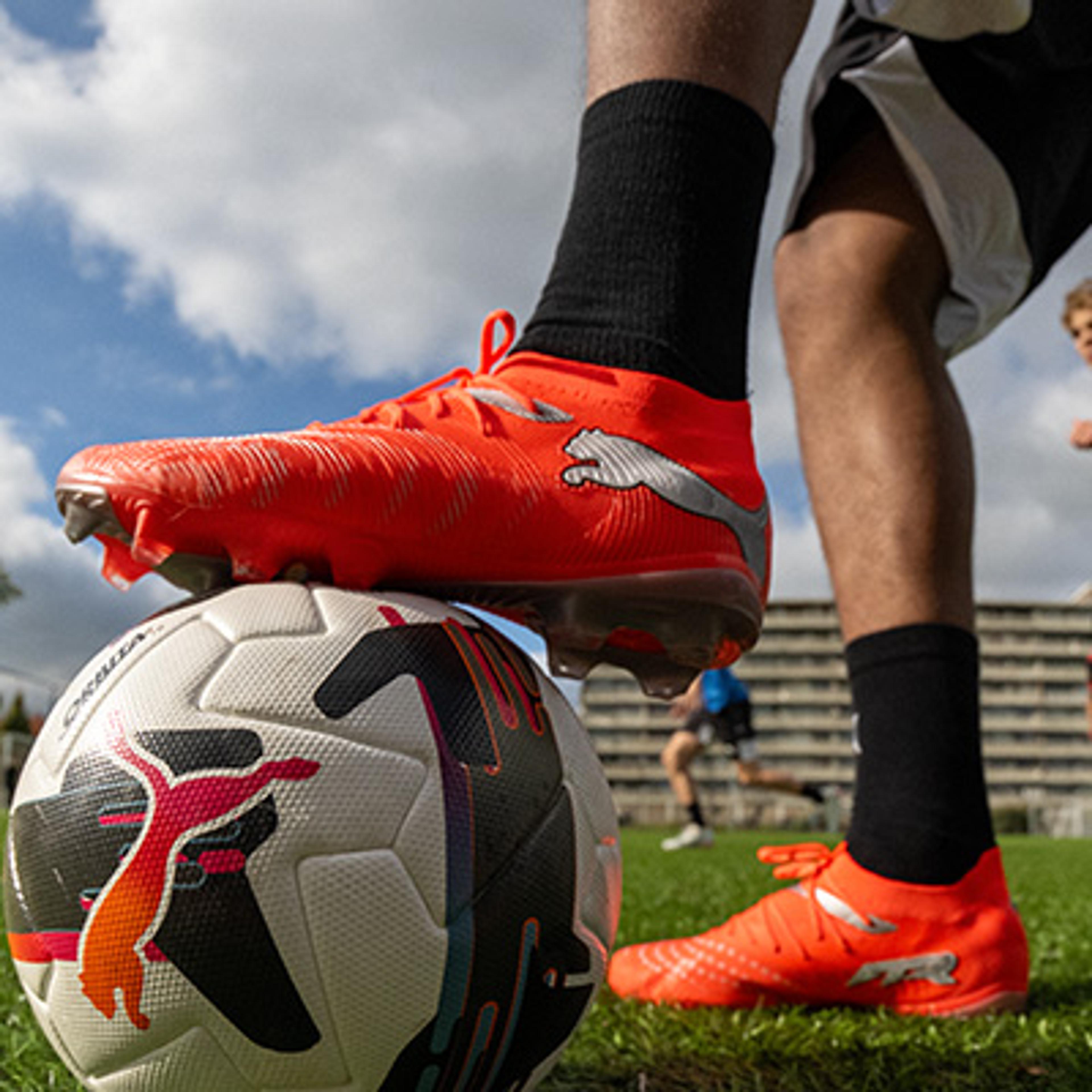A soccer player's foot in a bright orange Puma cleat rests on a soccer ball on a grassy field.