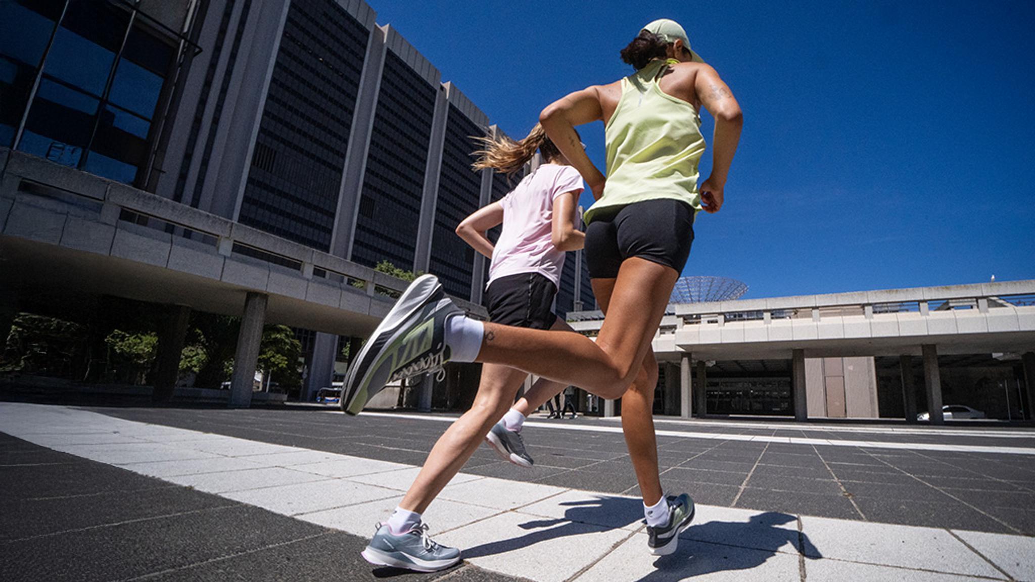 Two women running from behind on a paved urban path with modern buildings and a clear blue sky.
