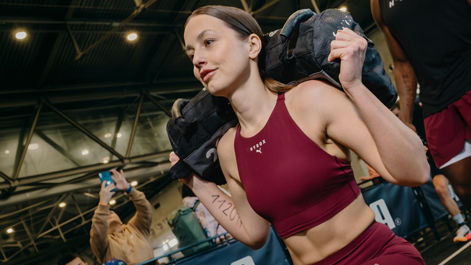A strong woman in dark red athletic wear carries a black sandbag on her shoulders.