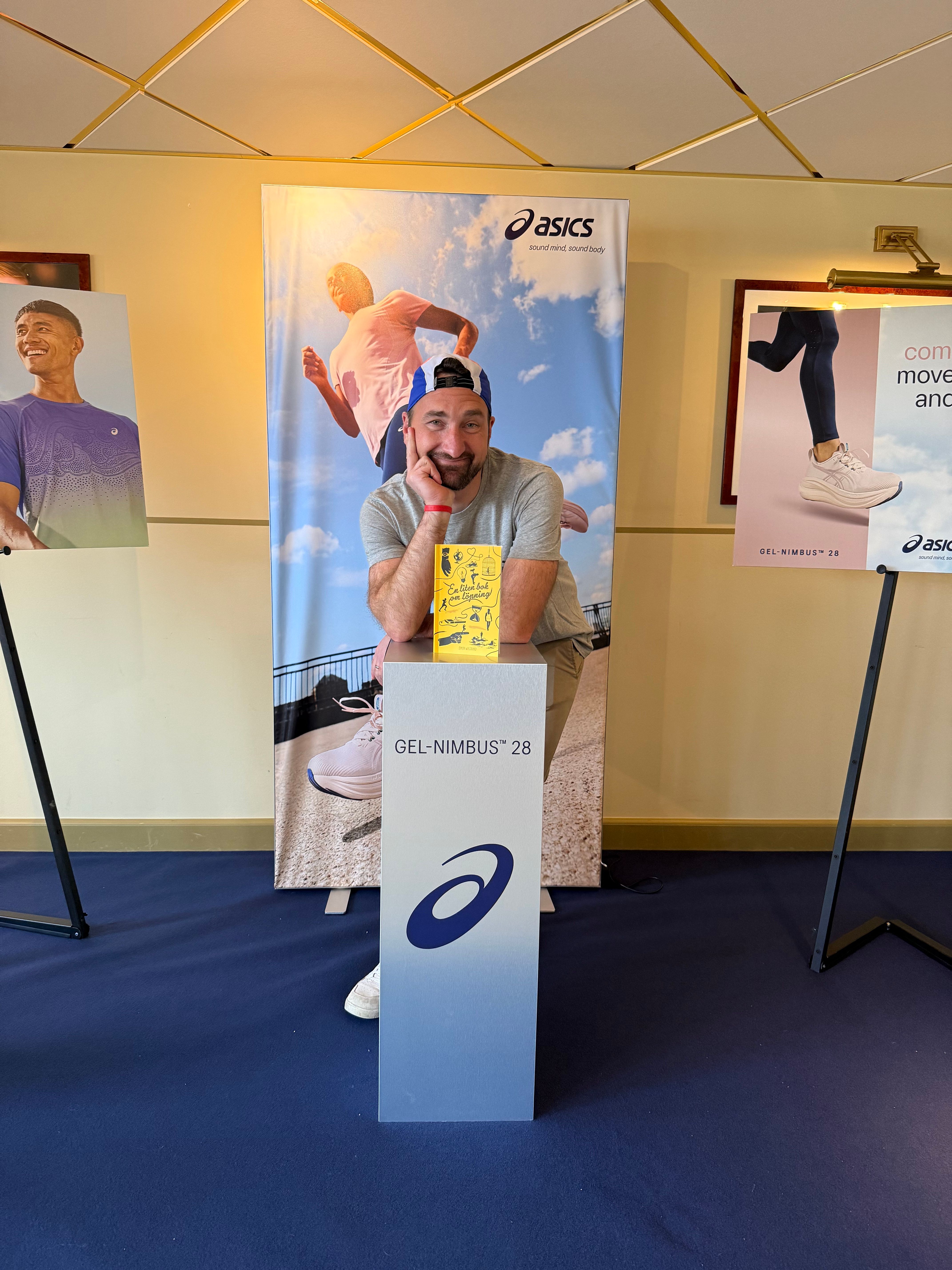 A smiling man leans on an Asics "GEL-NIMBUS 28" display stand, surrounded by Asics brand banners.