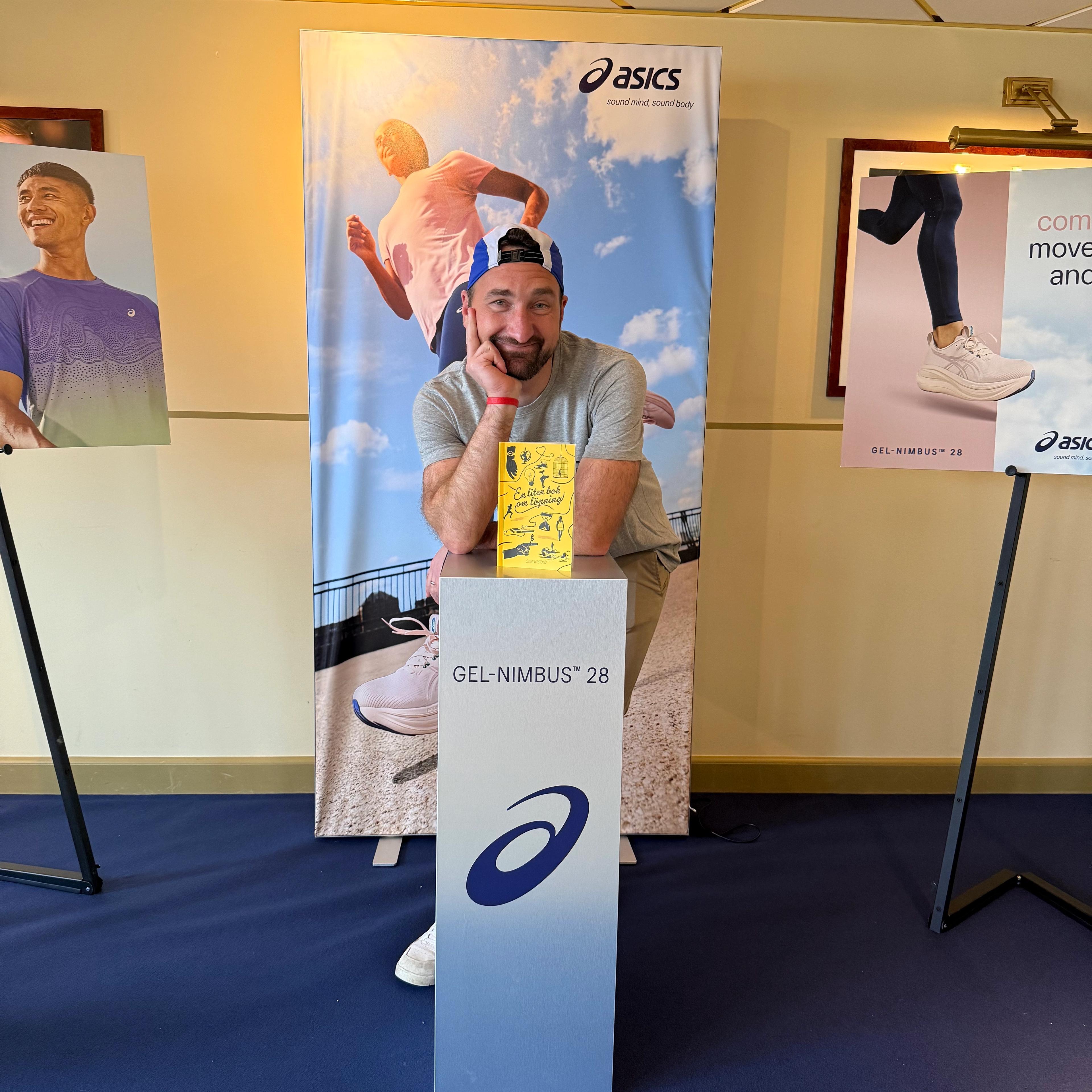 A smiling man leans on an Asics "GEL-NIMBUS 28" display stand, surrounded by Asics brand banners.