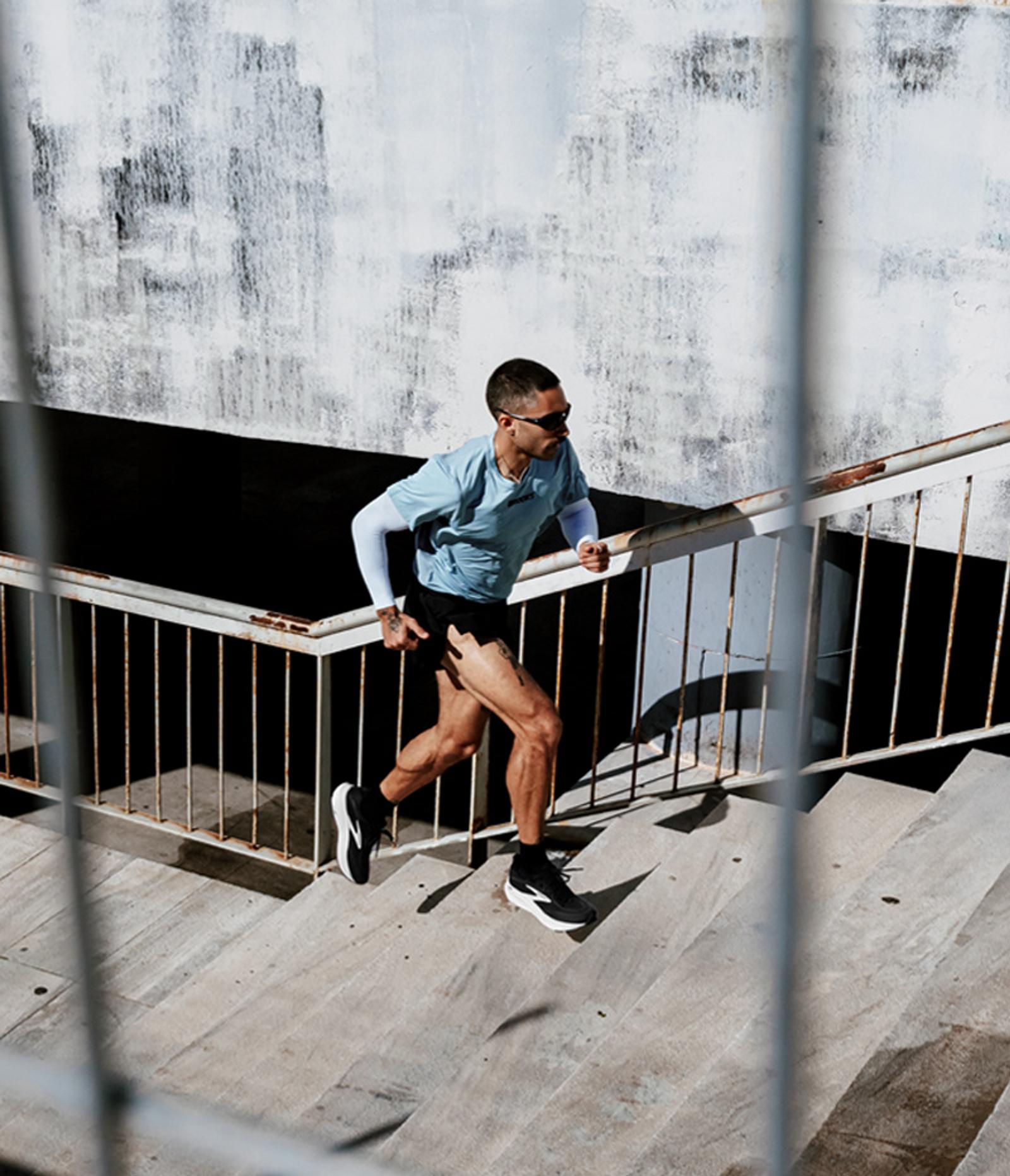 A man in athletic wear and sunglasses runs up concrete stairs with metal railings.
