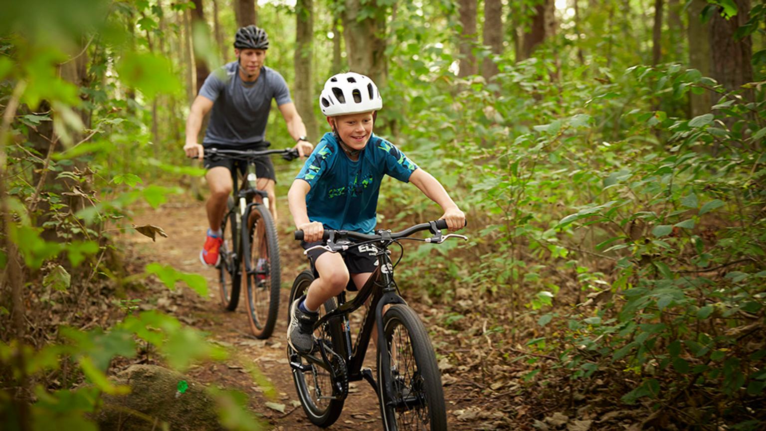 a man and a boy are riding bicycles down a trail in the woods .