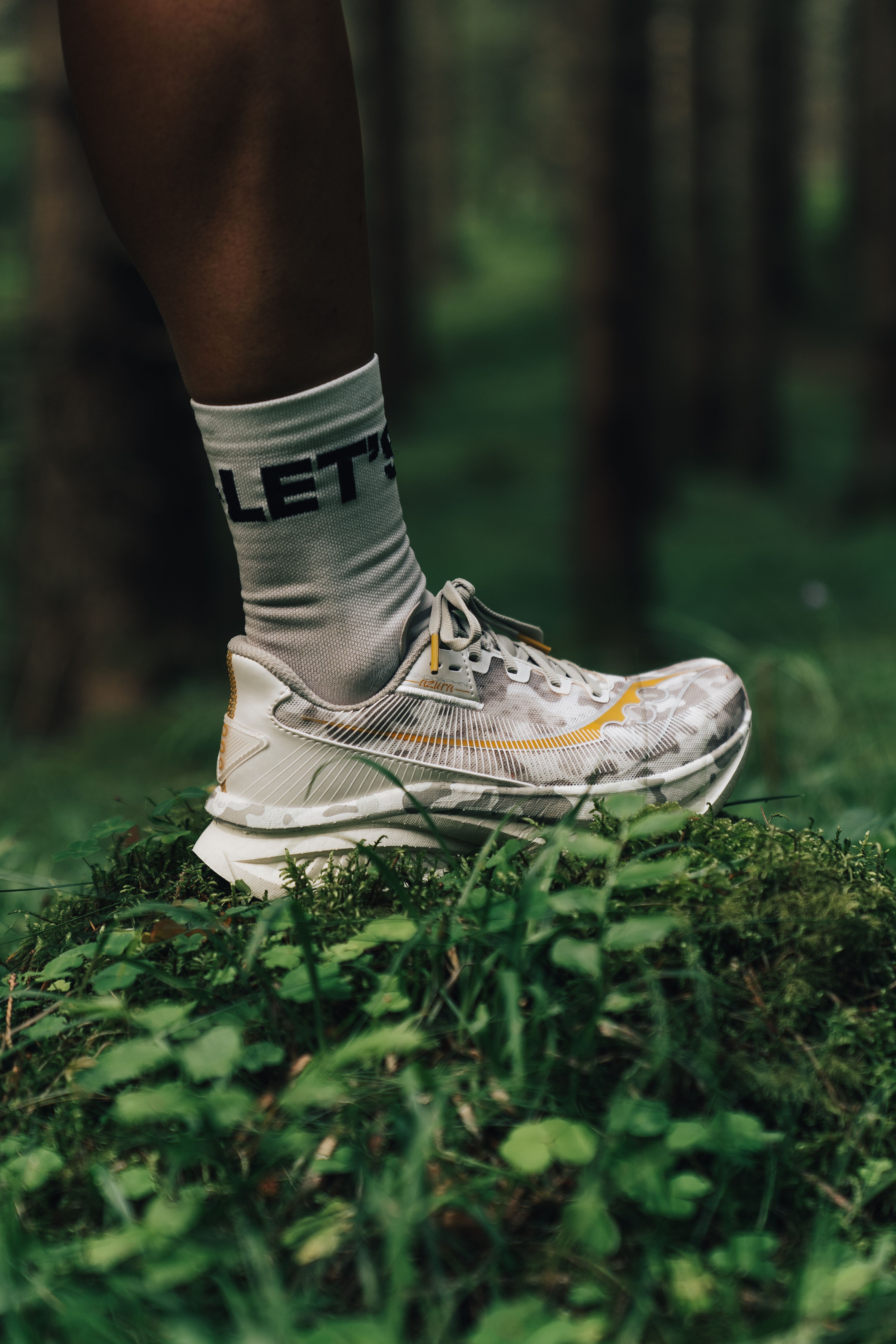 A foot in a grey camouflage running shoe with yellow accents and a "LET'S" sock, standing on moss in a forest.