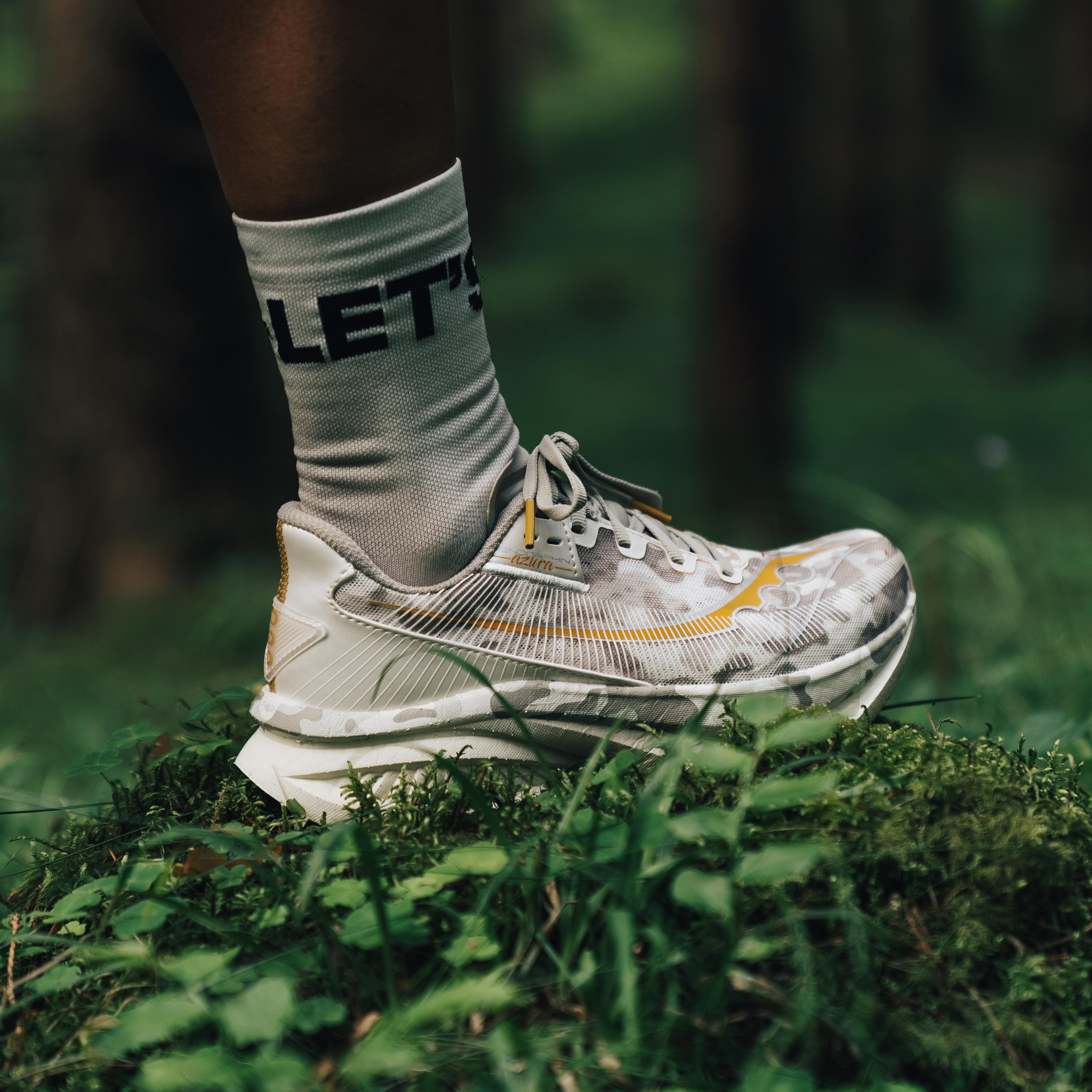 A leg and foot in a grey camo running shoe with yellow accents and a "LET'S" sock, on moss in a forest.