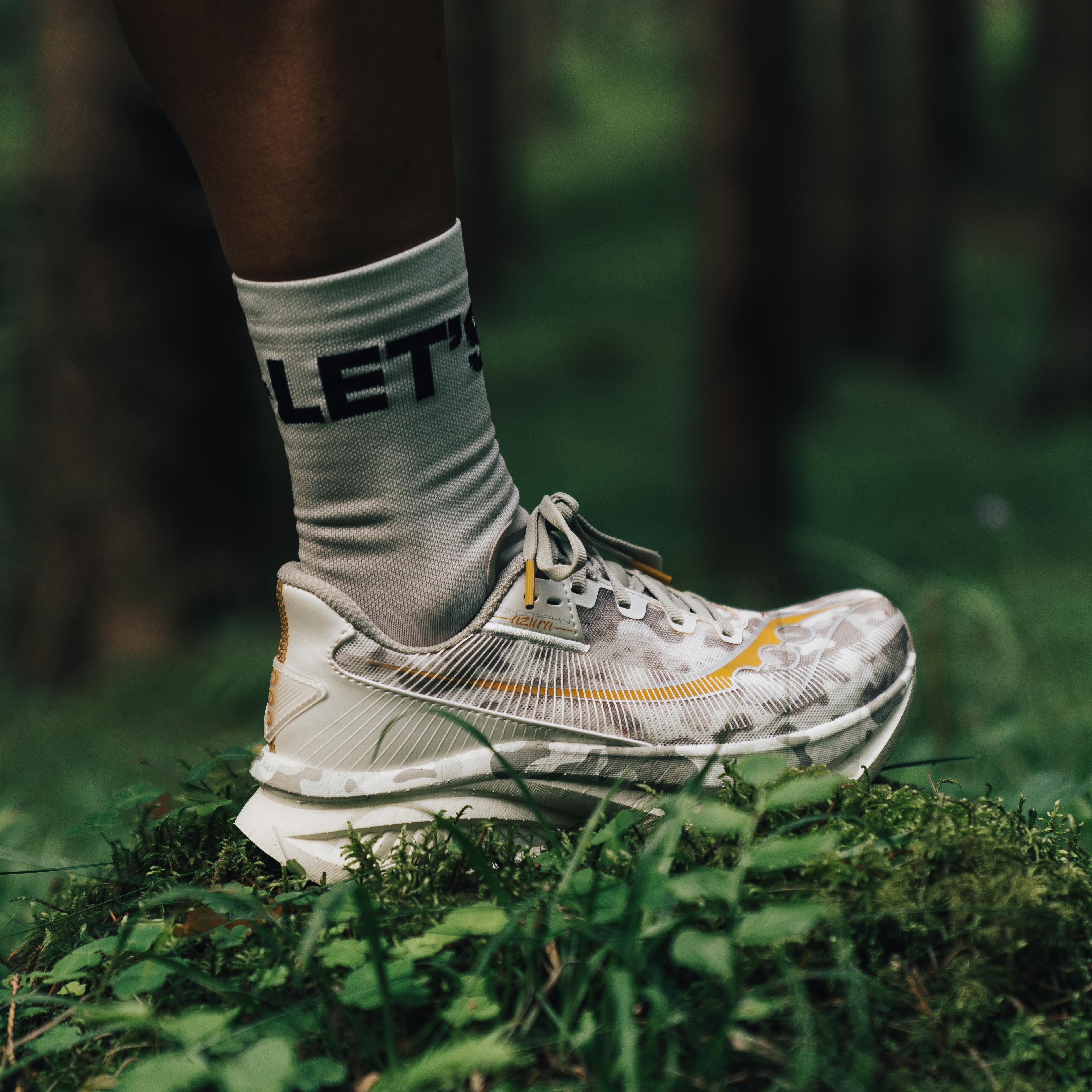 A foot in a grey camouflage running shoe with yellow accents and a "LET'S" sock, standing on moss in a forest.
