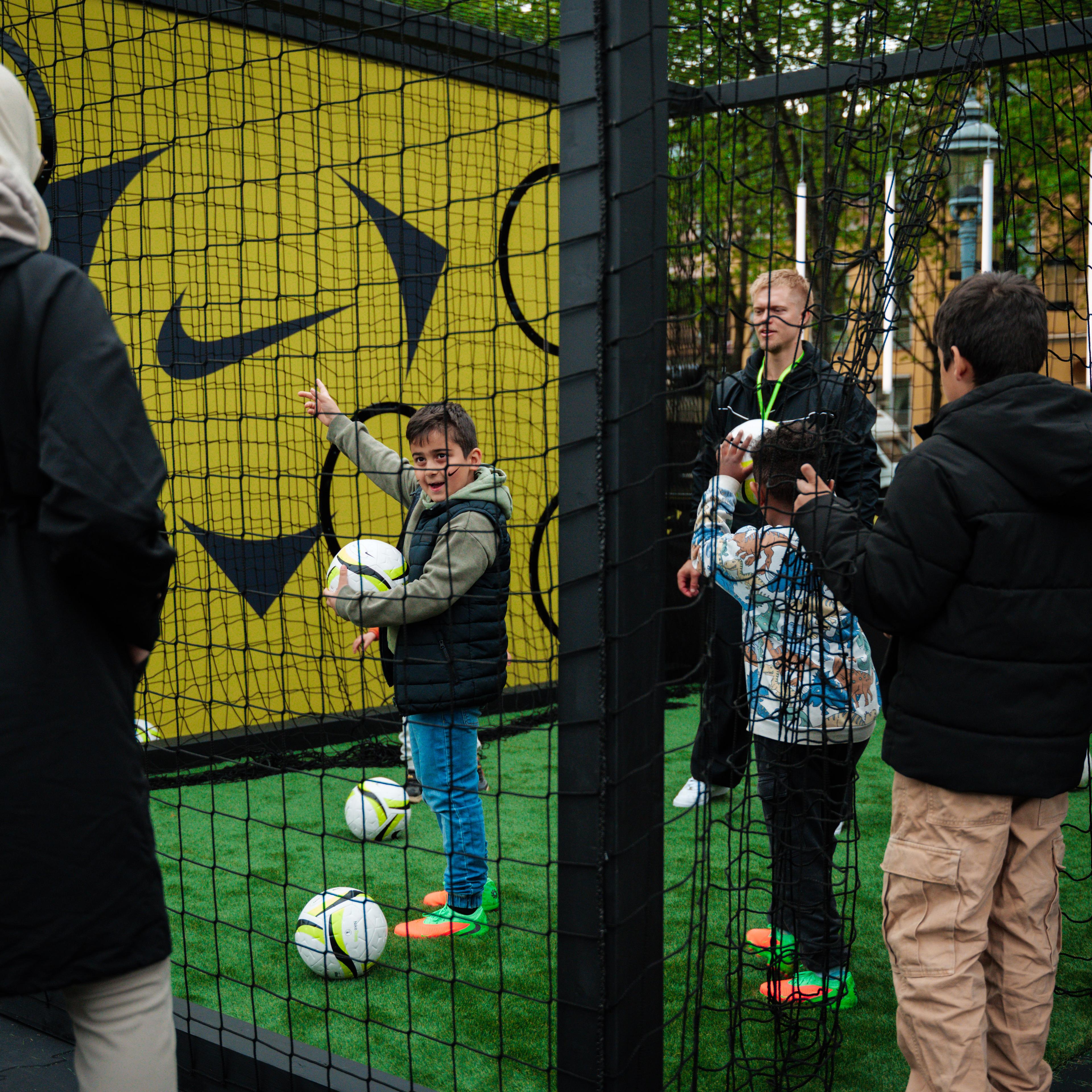 Children and adults playing soccer in a netted, Nike-branded turf area.