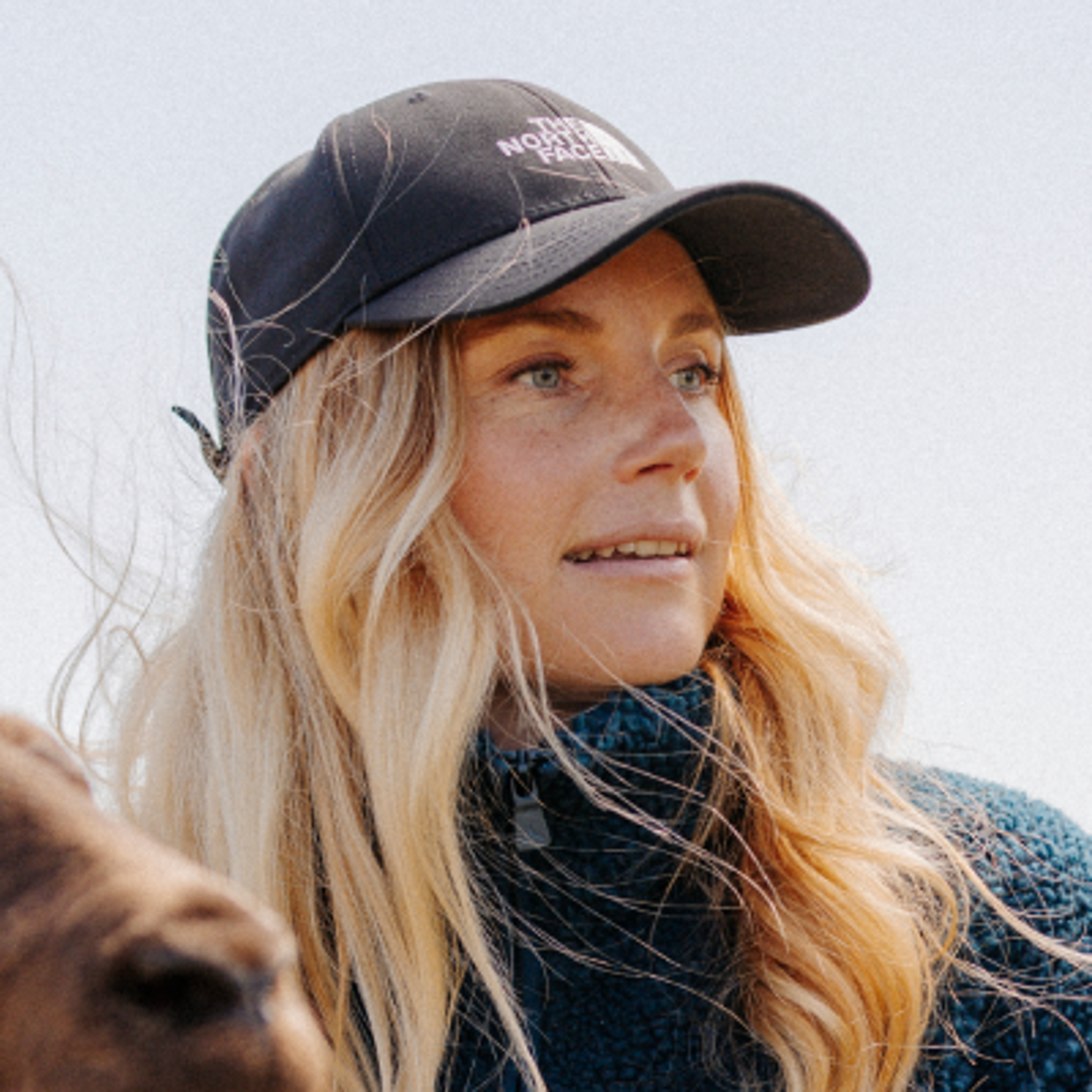 A woman in a North Face cap and quilted vest holds sunglasses, looking sideways with a slight smile.