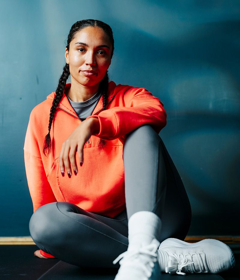 A woman with braids sits on the floor wearing an orange hoodie, grey leggings, and white sneakers, looking at the camera.