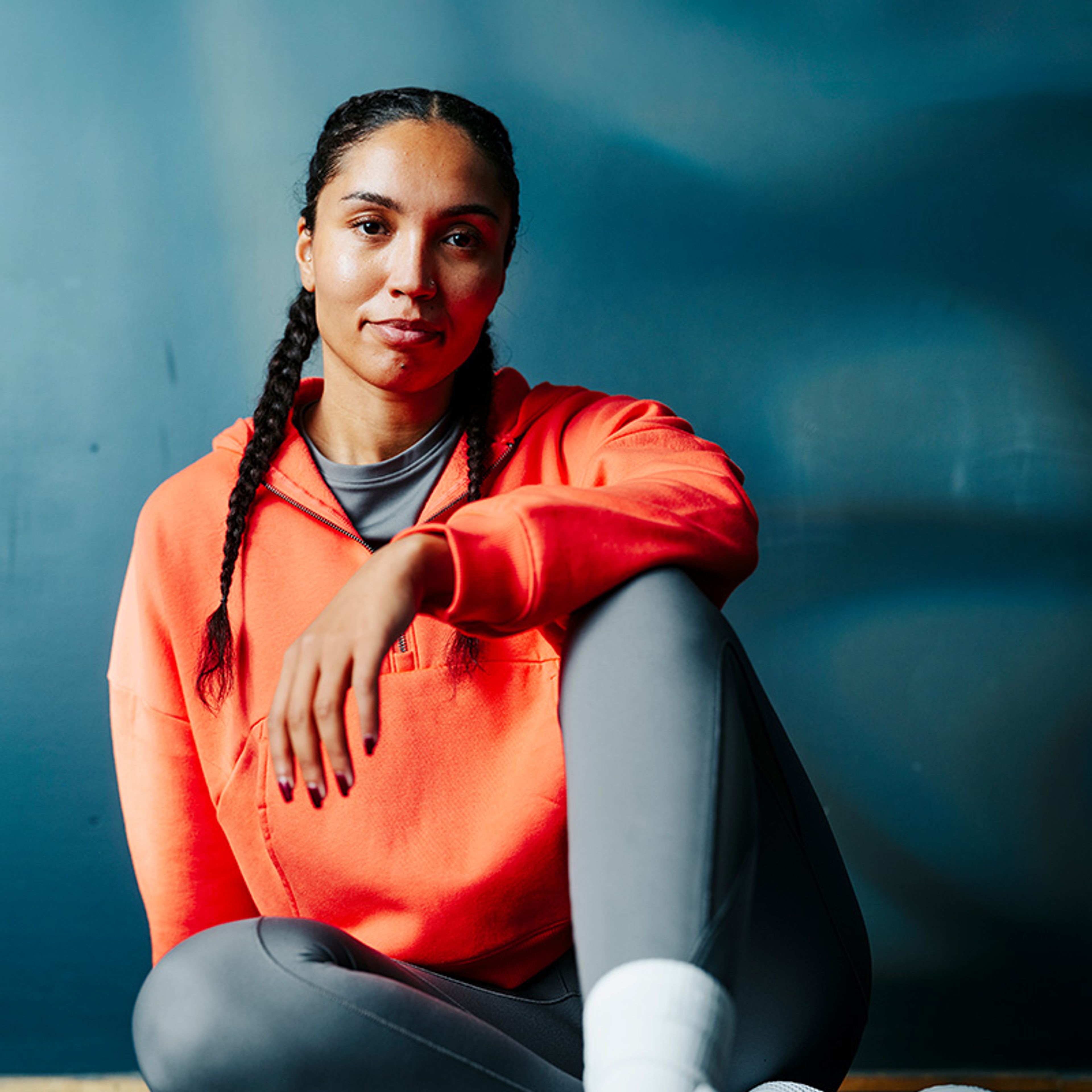 A woman with braids sits on the floor wearing an orange hoodie, grey leggings, and white sneakers, looking at the camera.