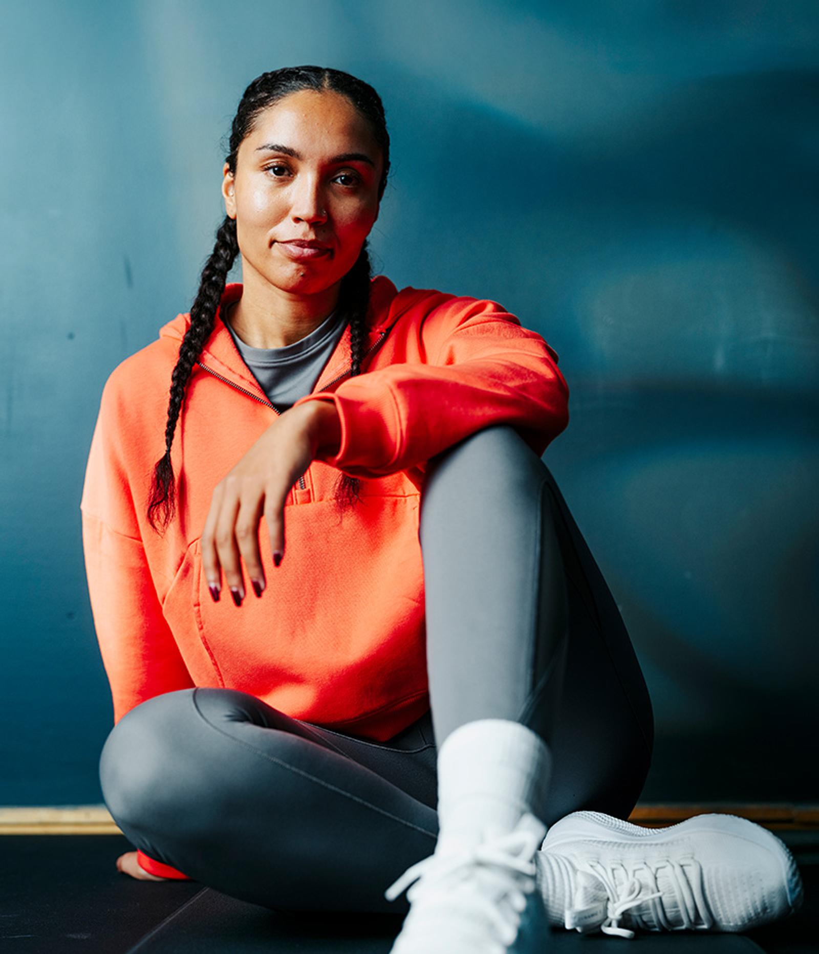 A person with braids sits on the floor wearing an orange hoodie, grey leggings, and white sneakers, looking at the camera.