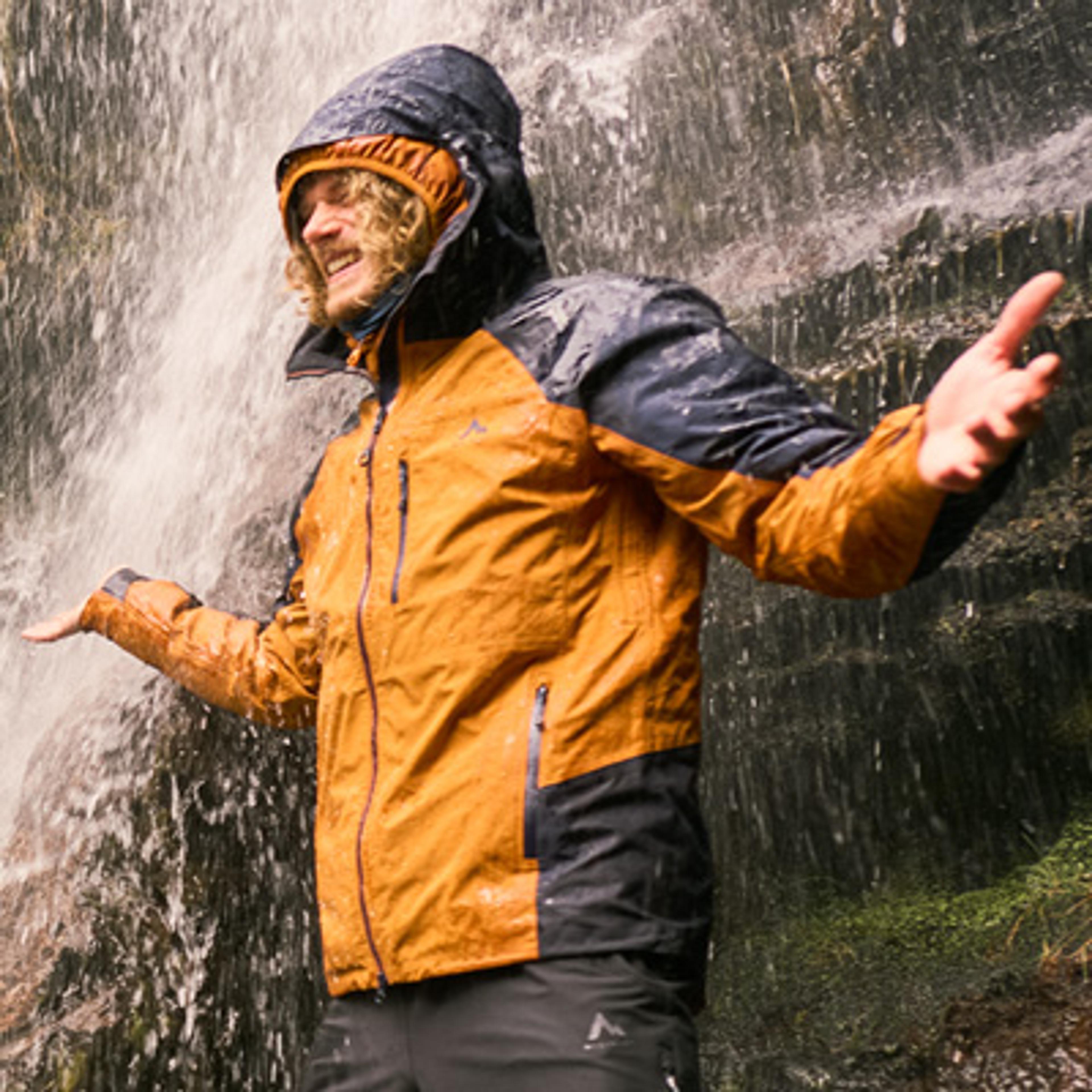 A person in a hooded orange and dark blue outdoor jacket smiles while standing in a waterfall, arms open.