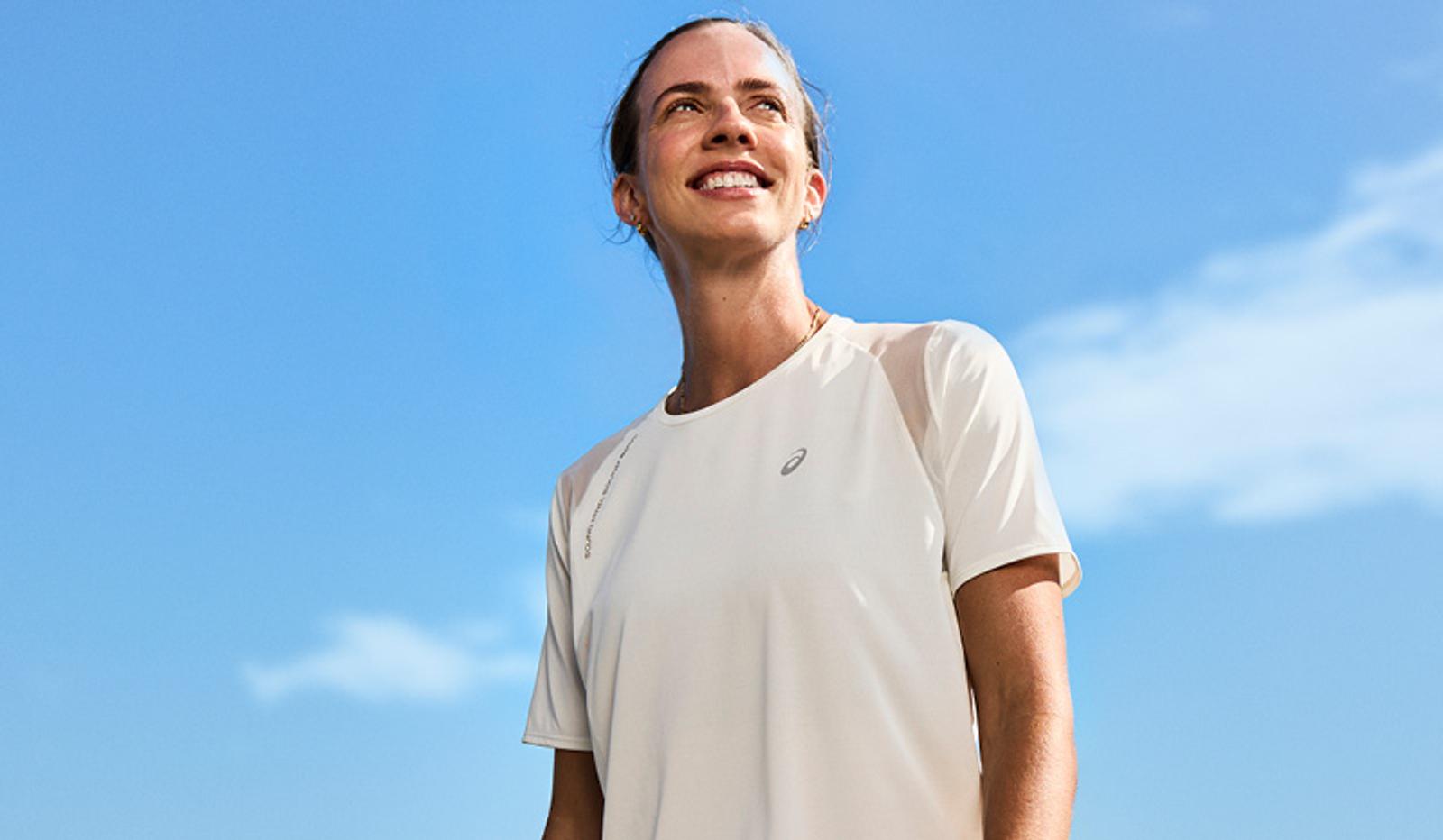 Smiling woman in white Asics shirt looking up, blue sky background.